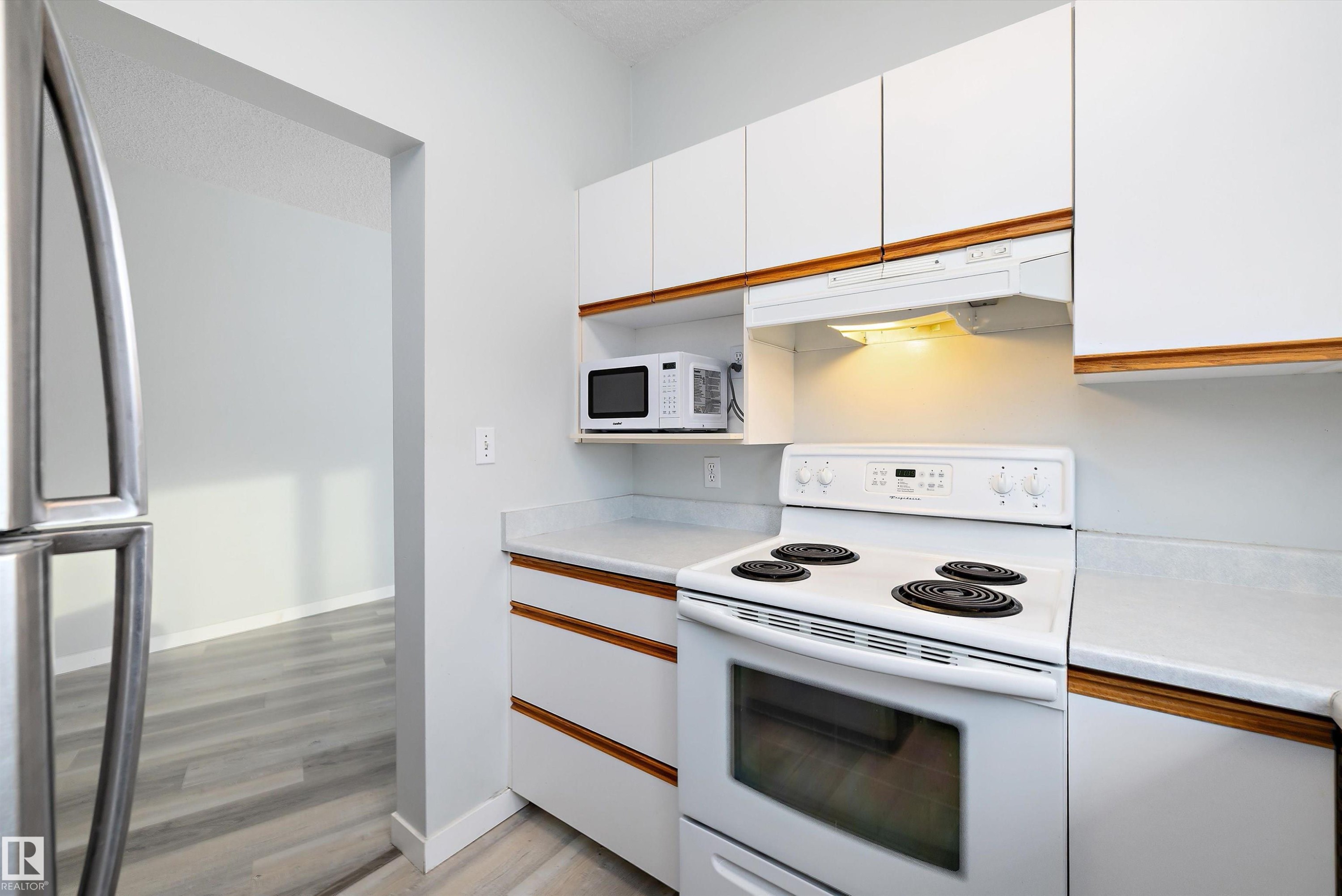 Kitchen with light countertops, white appliances, white cabinets, under cabinet range hood, and brown cabinetry - 205 10809 Saskatchewan Drive, Edmonton, AB - Indoor Photo Showing Kitchen