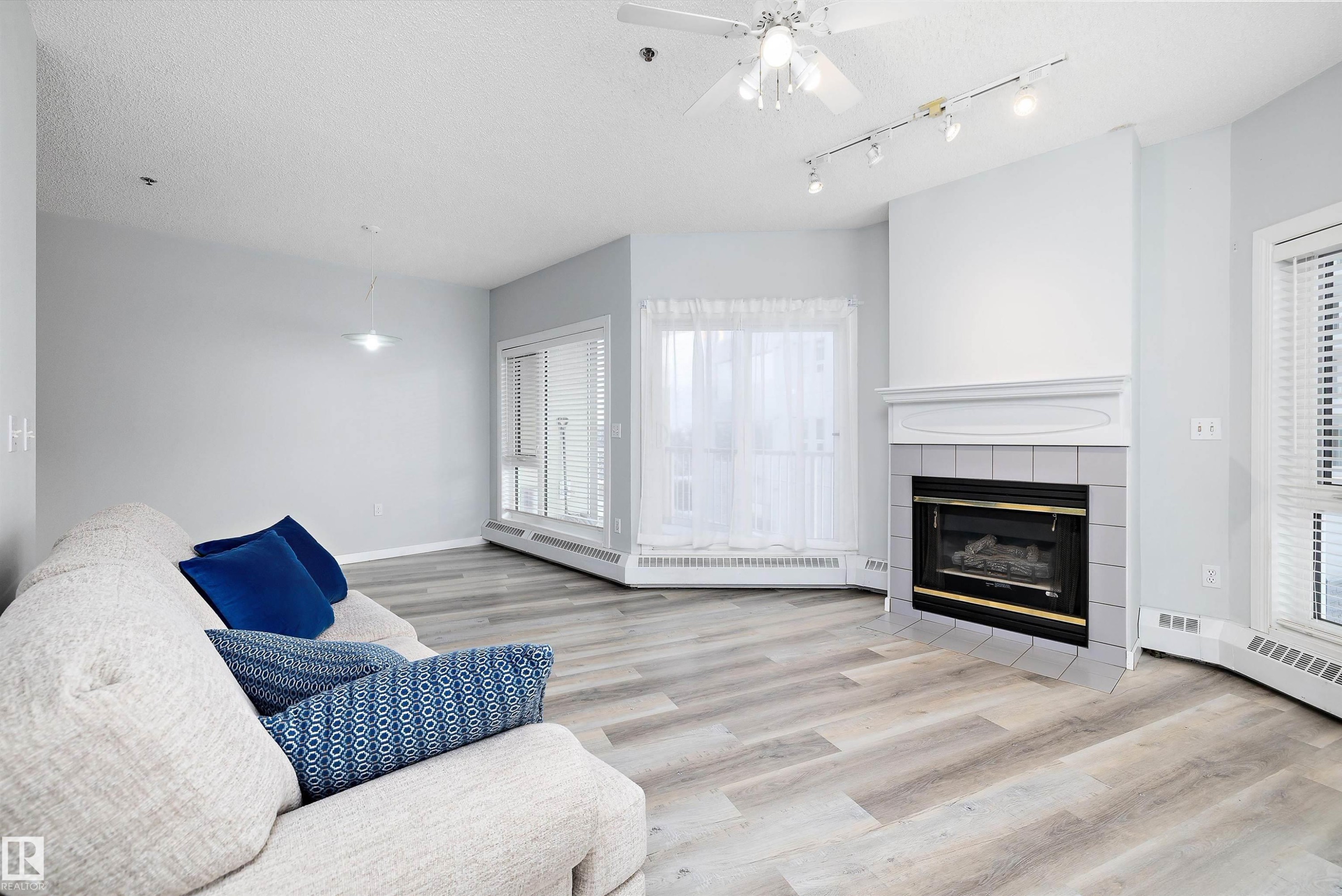 Living area featuring a fireplace, a textured ceiling, light wood finished floors, ceiling fan, and a baseboard radiator - 205 10809 Saskatchewan Drive, Edmonton, AB - Indoor Photo Showing Living Room With Fireplace