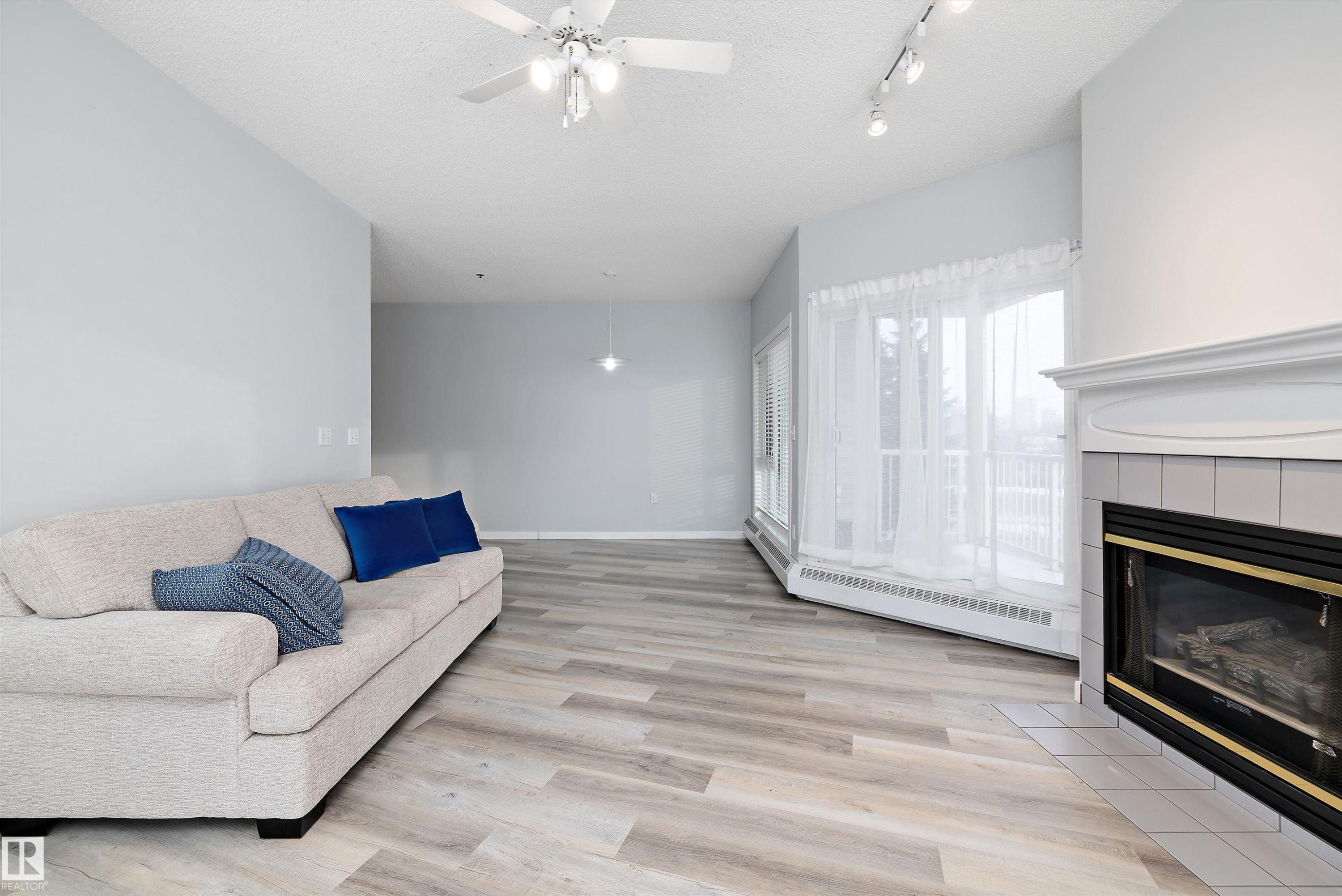 Living area with a textured ceiling, a ceiling fan, light wood-type flooring, a tile fireplace, and baseboard heating - 205 10809 Saskatchewan Drive, Edmonton, AB - Indoor Photo Showing Living Room With Fireplace