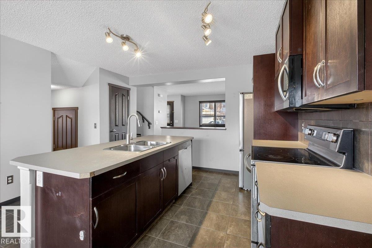 Kitchen featuring dark brown cabinetry, appliances with stainless steel finishes, light countertops, a textured ceiling, and an island with sink - 3623 13 Street, Edmonton, AB - Indoor Photo Showing Kitchen With Double Sink