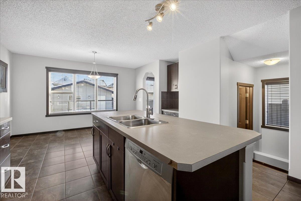 Kitchen with dark brown cabinets, stainless steel dishwasher, a kitchen island with sink, a textured ceiling, and dark tile patterned floors - 3623 13 Street, Edmonton, AB - Indoor Photo Showing Kitchen With Double Sink