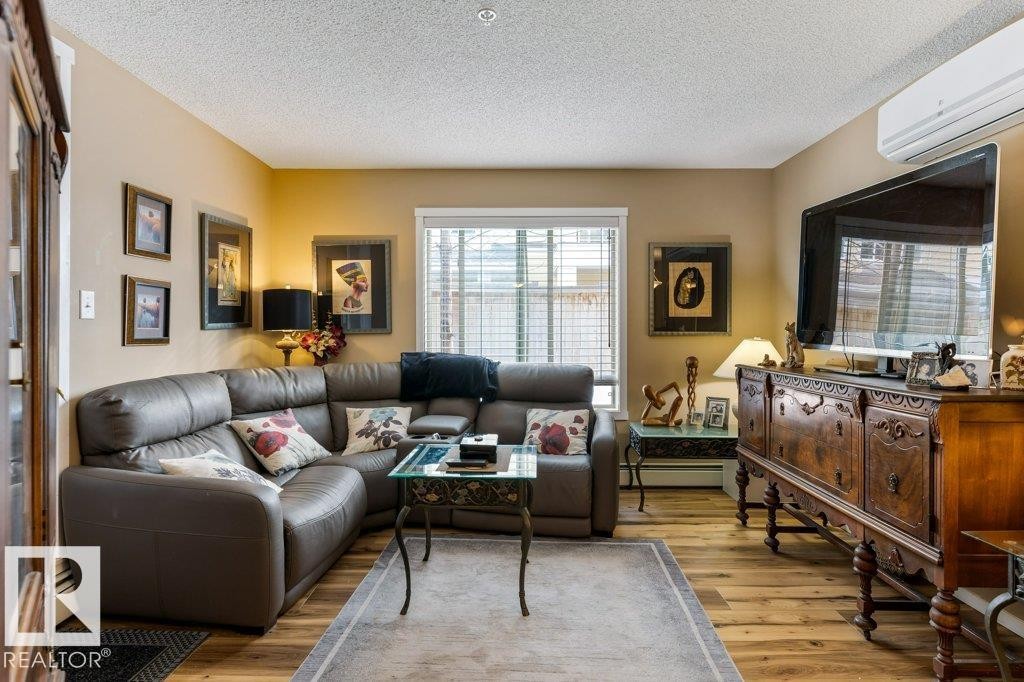 Living area with an AC wall unit, a textured ceiling, wood finished floors, and a baseboard heating unit - 107 25 Element Dr, St. Albert, AB - Indoor Photo Showing Living Room