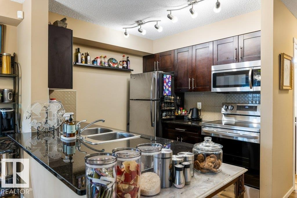 Kitchen featuring appliances with stainless steel finishes, dark stone counters, dark brown cabinets, backsplash, and a peninsula - 107 25 Element Dr, St. Albert, AB - Indoor Photo Showing Kitchen With Double Sink