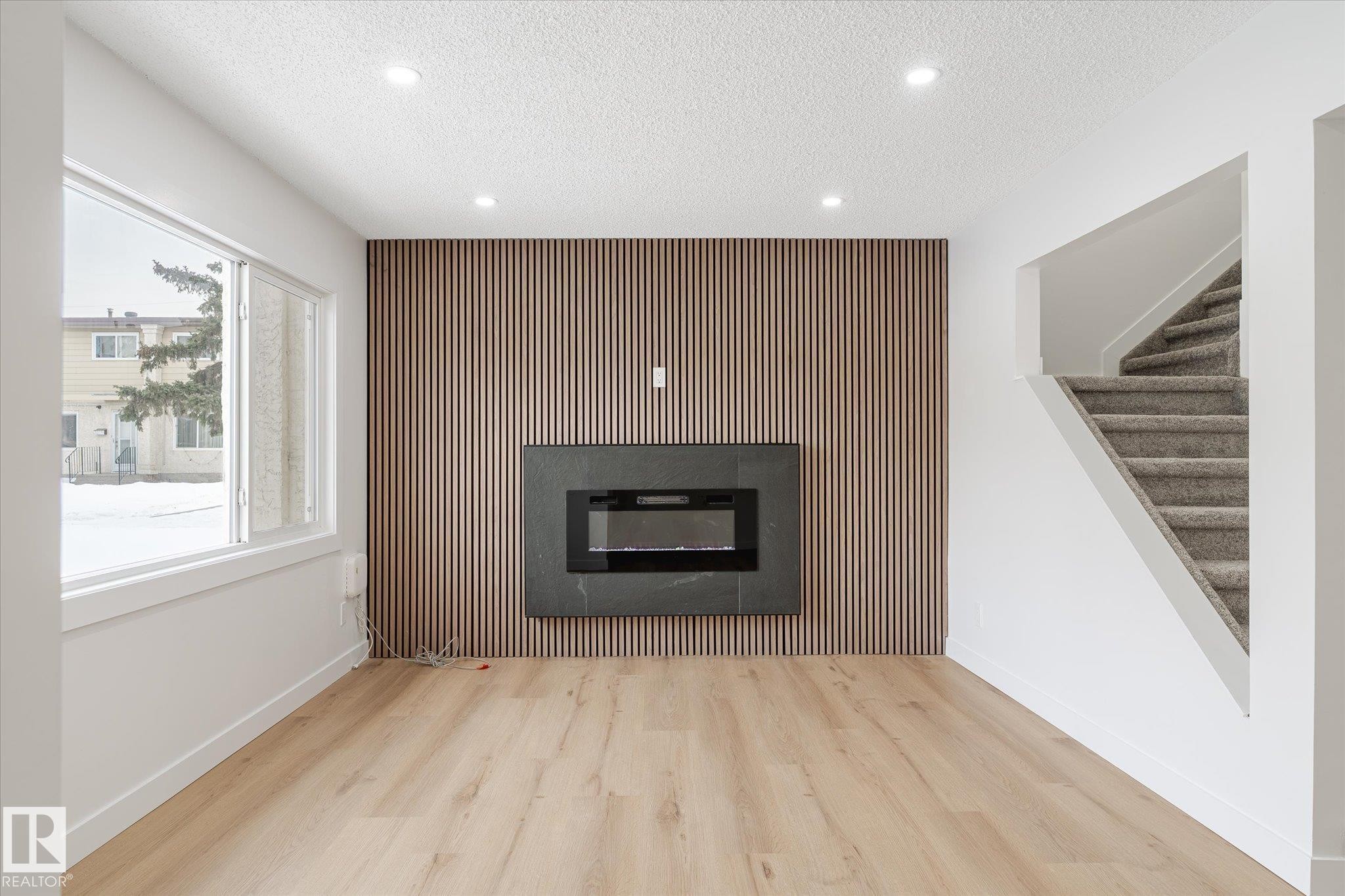 Unfurnished living room with an accent wall, a glass covered fireplace, wood finished floors, a textured ceiling, and stairs - 5105 106A Street, Edmonton, AB - Indoor Photo Showing Living Room With Fireplace