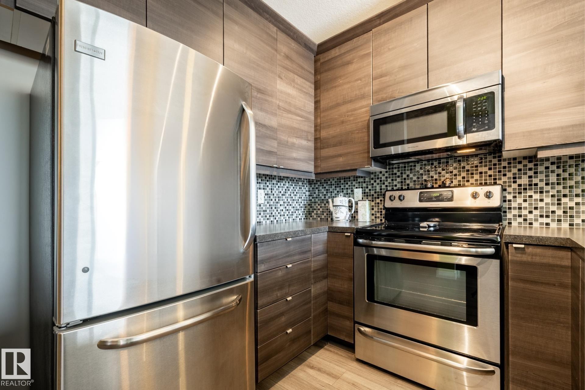 Kitchen featuring stainless steel appliances, dark countertops, decorative backsplash, light wood-style floors, and modern cabinets - 407 2590 Anderson Way, Edmonton, AB - Indoor Photo Showing Kitchen