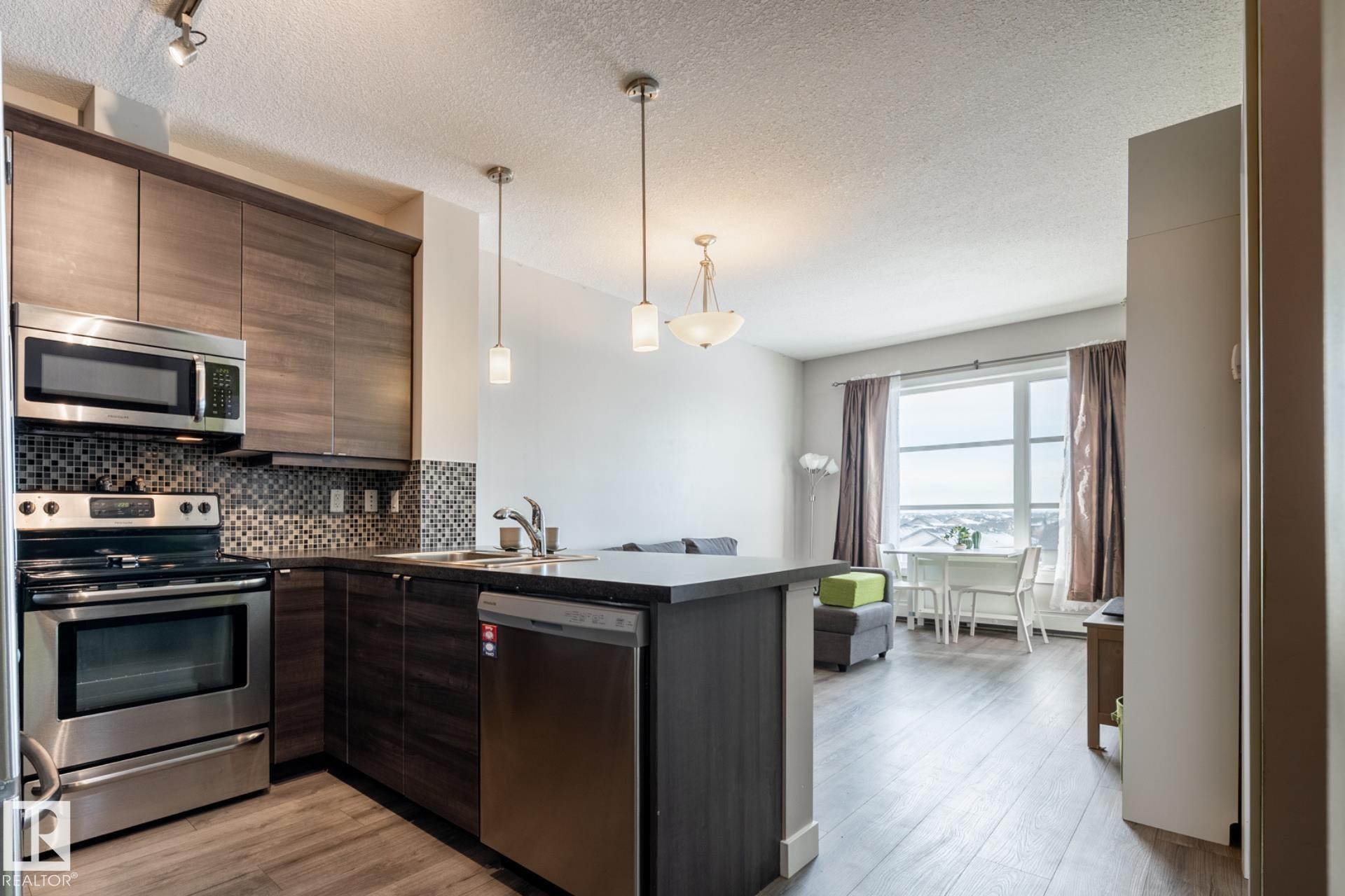 Kitchen featuring dark countertops, stainless steel appliances, a peninsula, dark wood finish cabinets, and light wood-type flooring - 407 2590 Anderson Way, Edmonton, AB - Indoor Photo Showing Kitchen