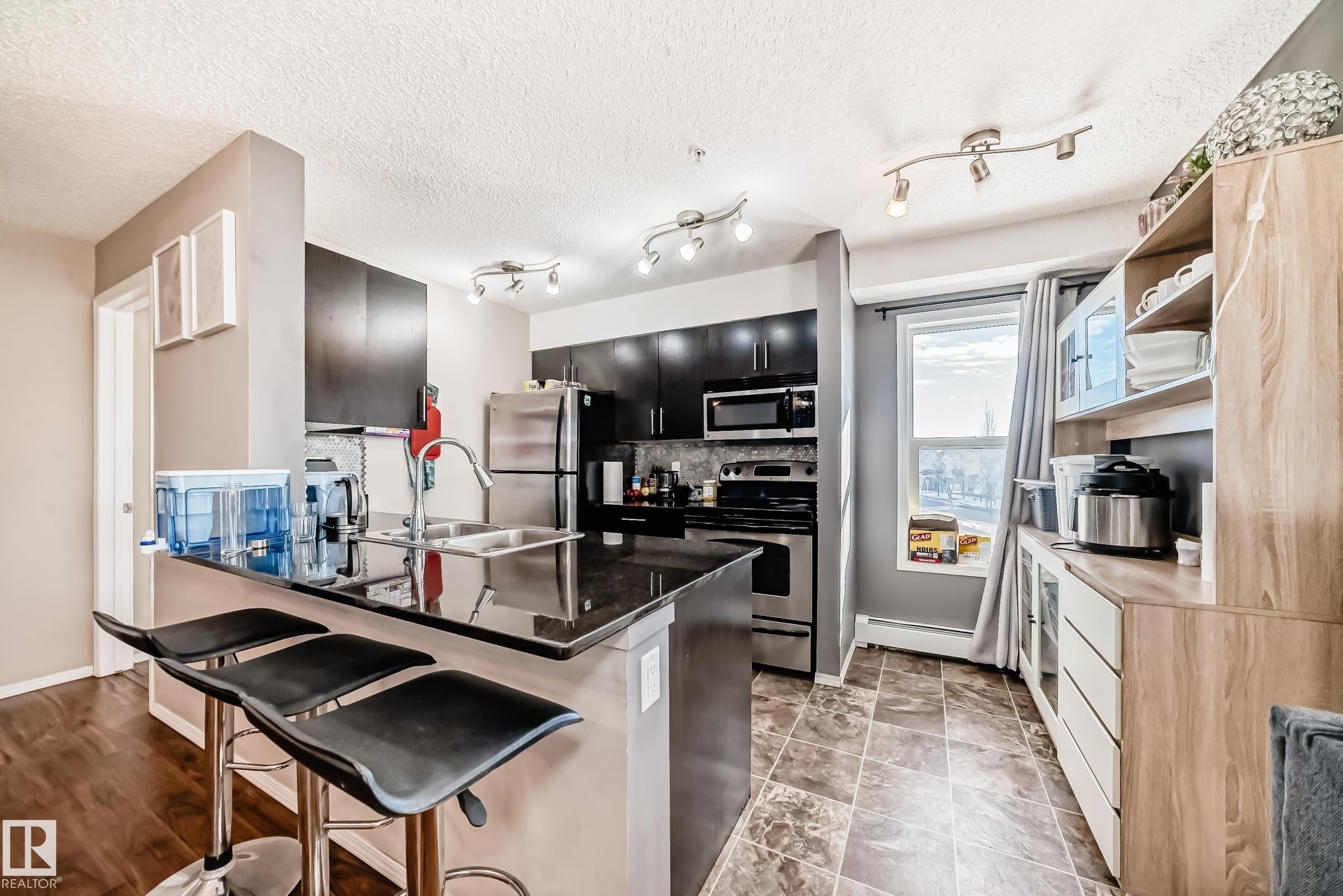 Kitchen featuring dark cabinets, stainless steel appliances, a breakfast bar area, dark stone counters, and a baseboard radiator - 302 530 Watt Boulevard Sw, Edmonton, AB - Indoor Photo Showing Kitchen With Stainless Steel Kitchen With Double Sink