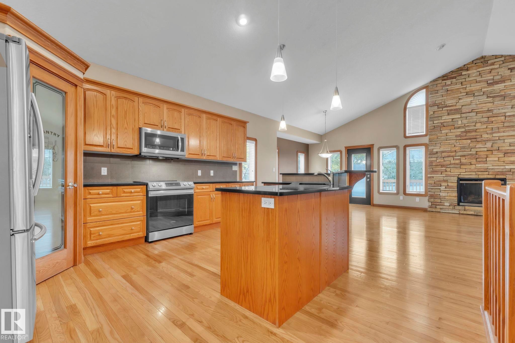 Kitchen with hanging light fixtures, decorative backsplash, stainless steel appliances, a stone fireplace, and light wood finished floors - 1217 Summerside Drive, Edmonton, AB - Indoor Photo Showing Kitchen