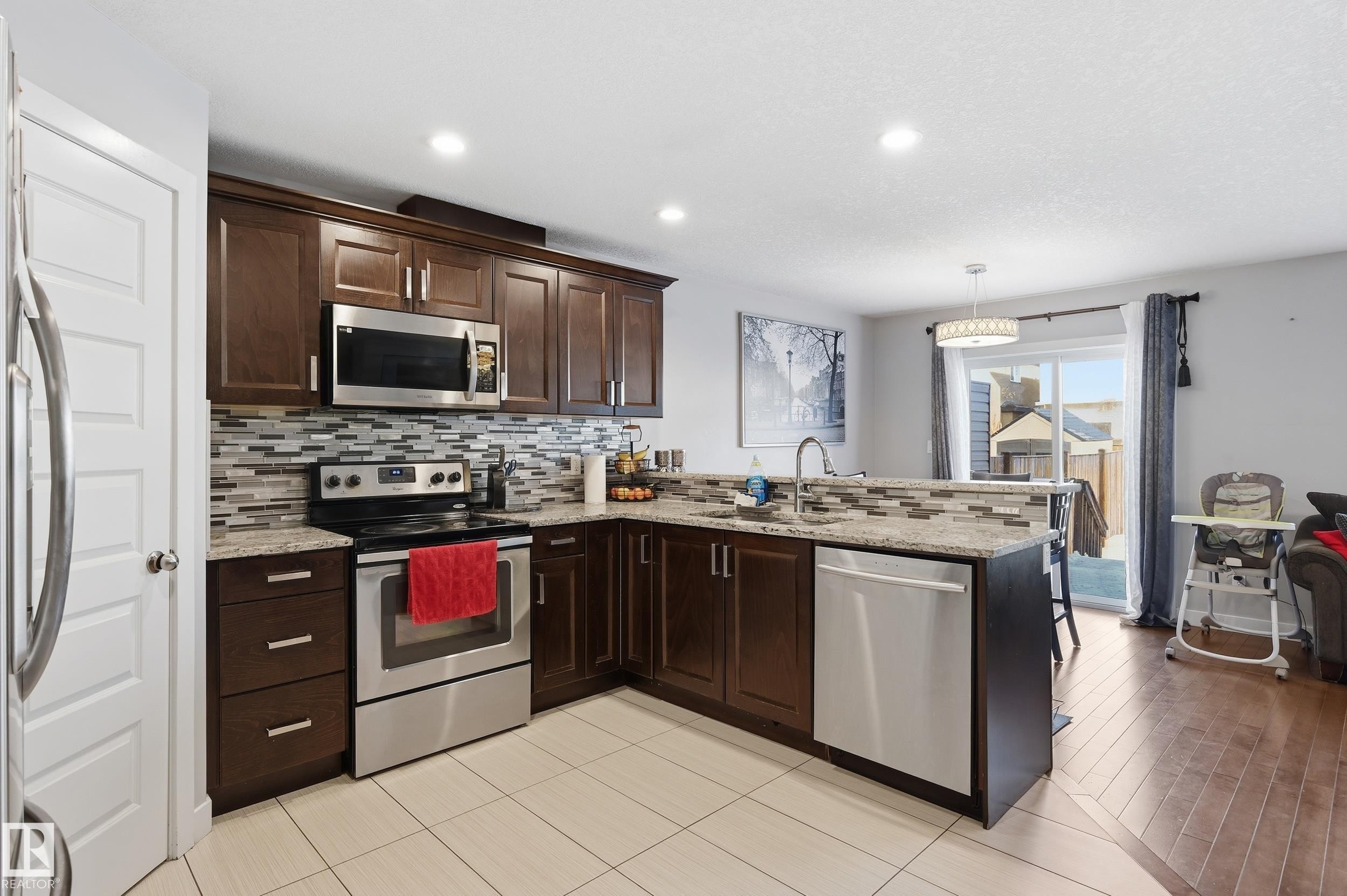 Kitchen featuring stainless steel appliances, tasteful backsplash, dark wood finish cabinets, light stone countertops, and a peninsula - 5978 167C Avenue, Edmonton, AB - Indoor Photo Showing Kitchen With Upgraded Kitchen