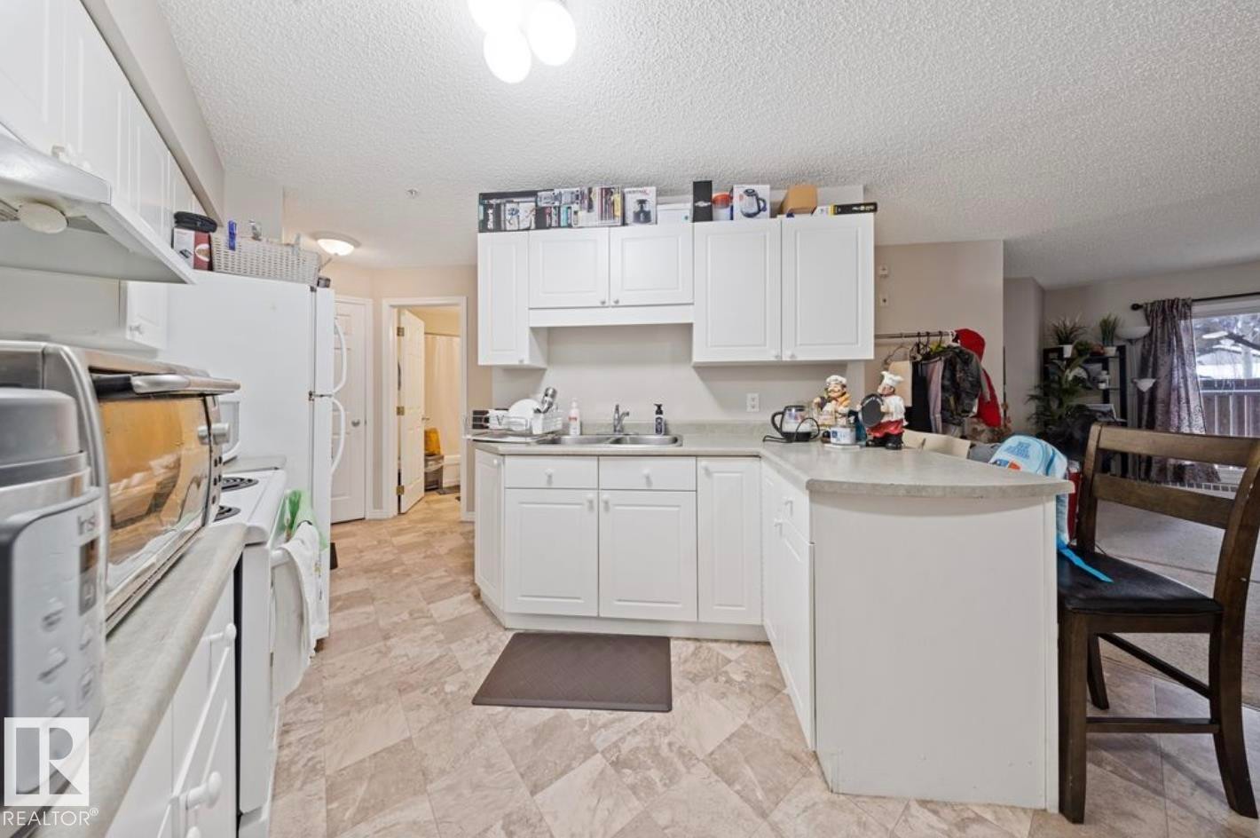 Kitchen featuring a peninsula, light countertops, white cabinetry, a textured ceiling, and white appliances - 103 12110 119 Ave, Edmonton, AB - Indoor Photo Showing Kitchen With Double Sink