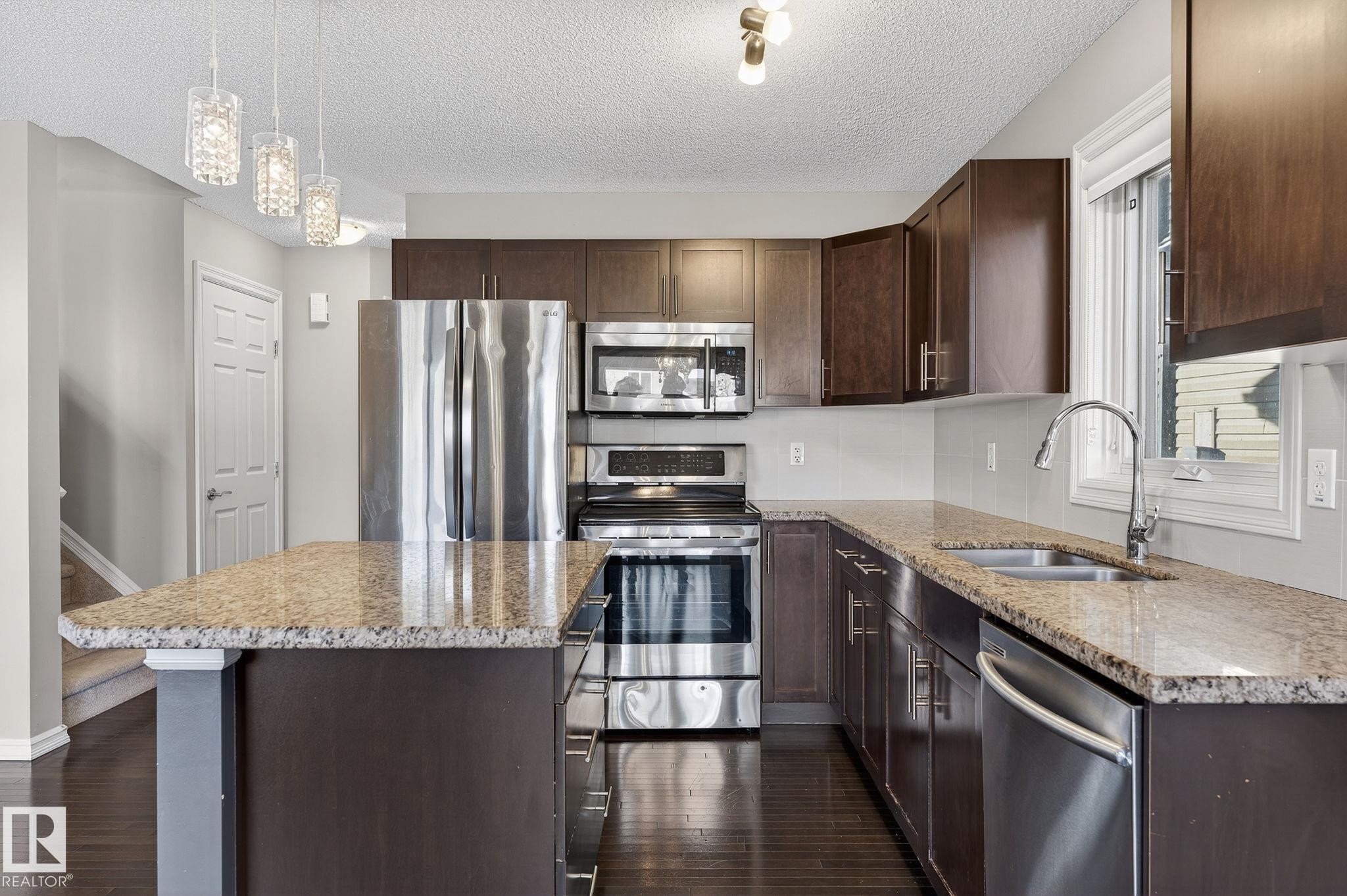 Kitchen with dark wood finish cabinets, stainless steel appliances, light stone countertops, and a textured ceiling - 2338 Casselman Crescent, Edmonton, AB - Indoor Photo Showing Kitchen With Stainless Steel Kitchen With Double Sink With Upgraded Kitchen
