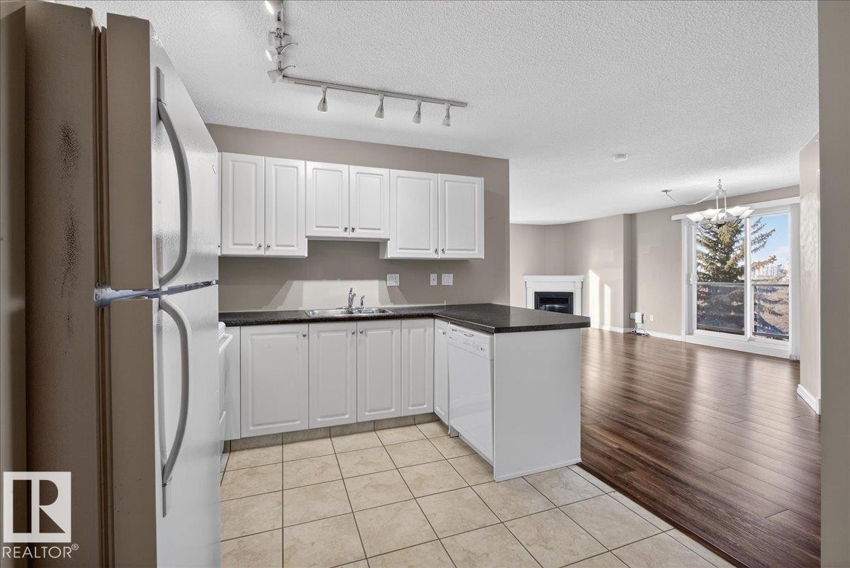 Kitchen with white appliances, white cabinets, a fireplace, a peninsula, and light tile patterned floors - 402 10649 Saskatchewan Drive, Edmonton, AB - Indoor Photo Showing Kitchen With Double Sink