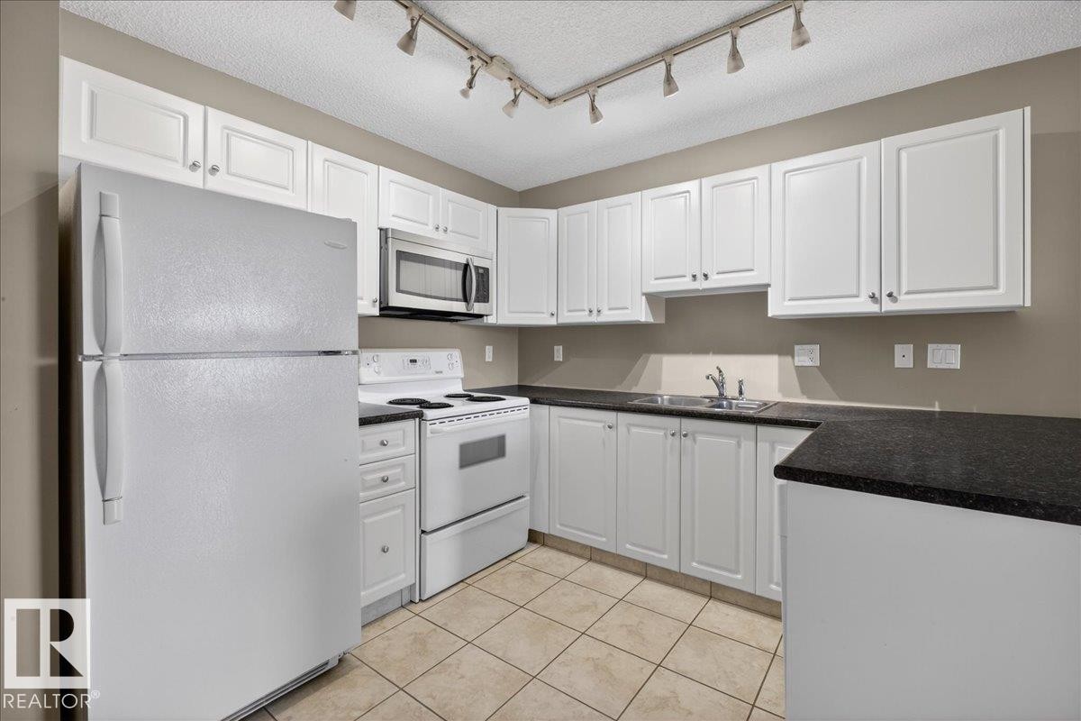 Kitchen featuring white appliances, white cabinetry, rail lighting, light tile patterned flooring, and a textured ceiling - 402 10649 Saskatchewan Drive, Edmonton, AB - Indoor Photo Showing Kitchen With Double Sink
