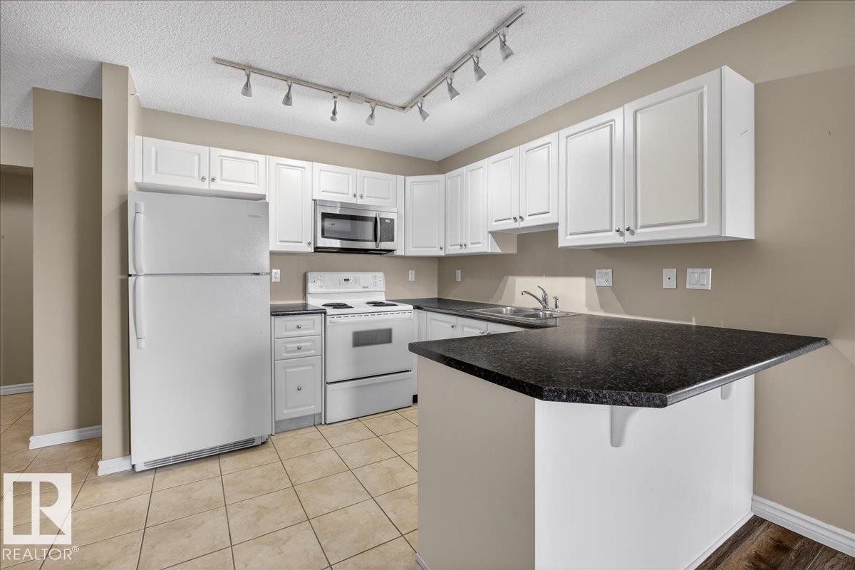 Kitchen featuring white appliances, white cabinets, rail lighting, dark countertops, and a peninsula - 402 10649 Saskatchewan Drive, Edmonton, AB - Indoor Photo Showing Kitchen With Double Sink