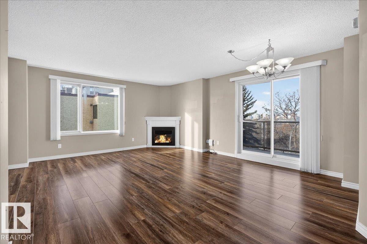 Unfurnished living room with a lit fireplace, dark wood-style flooring, a textured ceiling, and hanging lights - 402 10649 Saskatchewan Drive, Edmonton, AB - Indoor Photo Showing Living Room With Fireplace