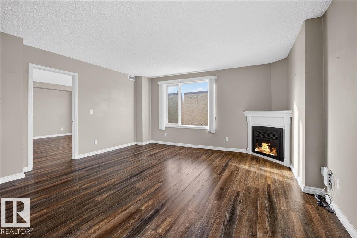 Unfurnished living room featuring dark wood-style flooring, a lit fireplace, and a textured ceiling - 402 10649 Saskatchewan Drive, Edmonton, AB - Indoor Photo Showing Other Room With Fireplace
