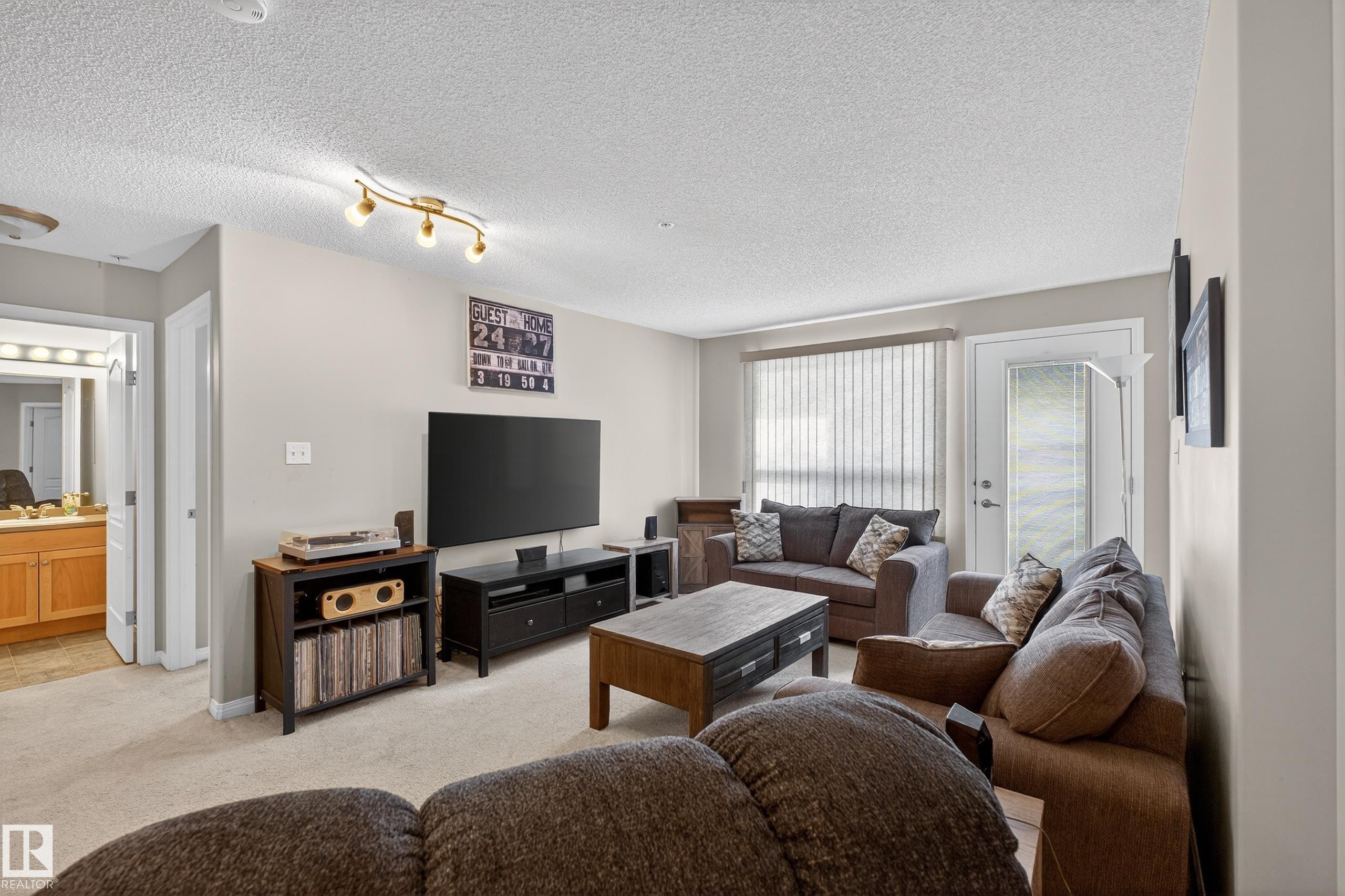 Carpeted living area featuring a textured ceiling and rail lighting - 131 1520 Hammond Gate, Edmonton, AB - Indoor Photo Showing Living Room