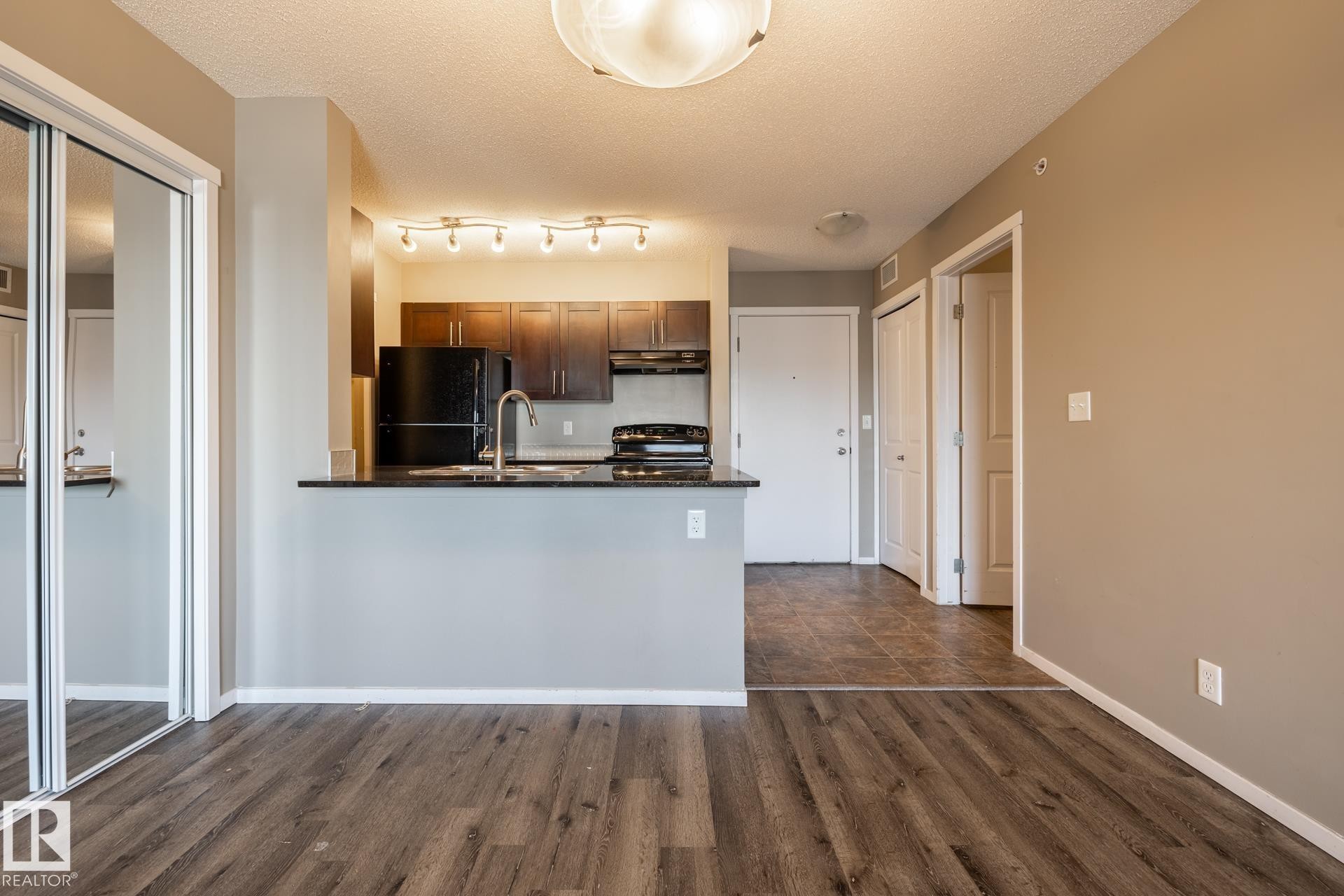 Kitchen featuring black appliances, a peninsula, a textured ceiling, dark wood-style floors, and dark stone counters - 433 3315 James Mowatt Trail, Edmonton, AB - Indoor Photo Showing Kitchen