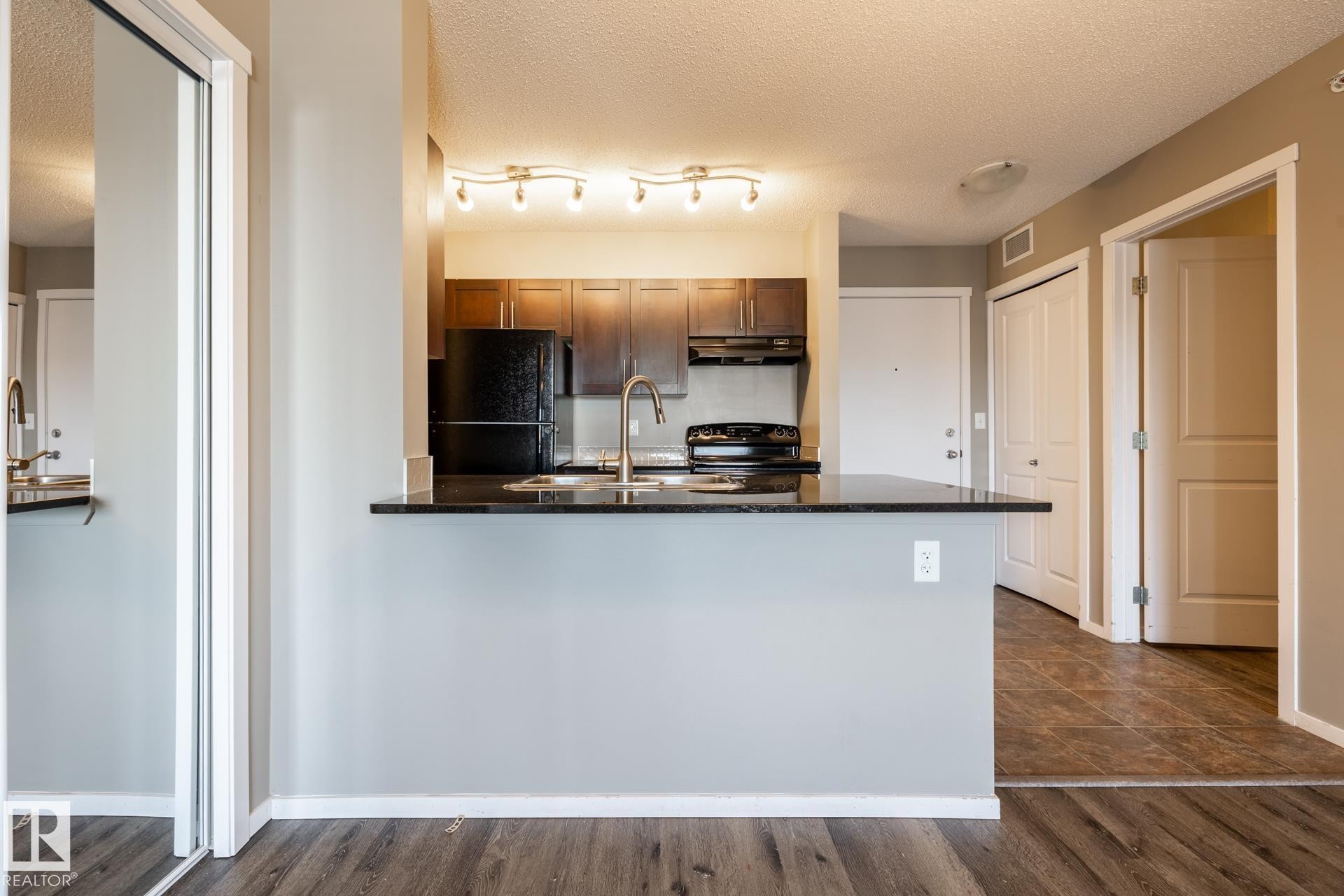 Kitchen featuring black appliances, a textured ceiling, a peninsula, dark stone countertops, and wood finish cabinetry - 433 3315 James Mowatt Trail, Edmonton, AB - Indoor Photo Showing Kitchen
