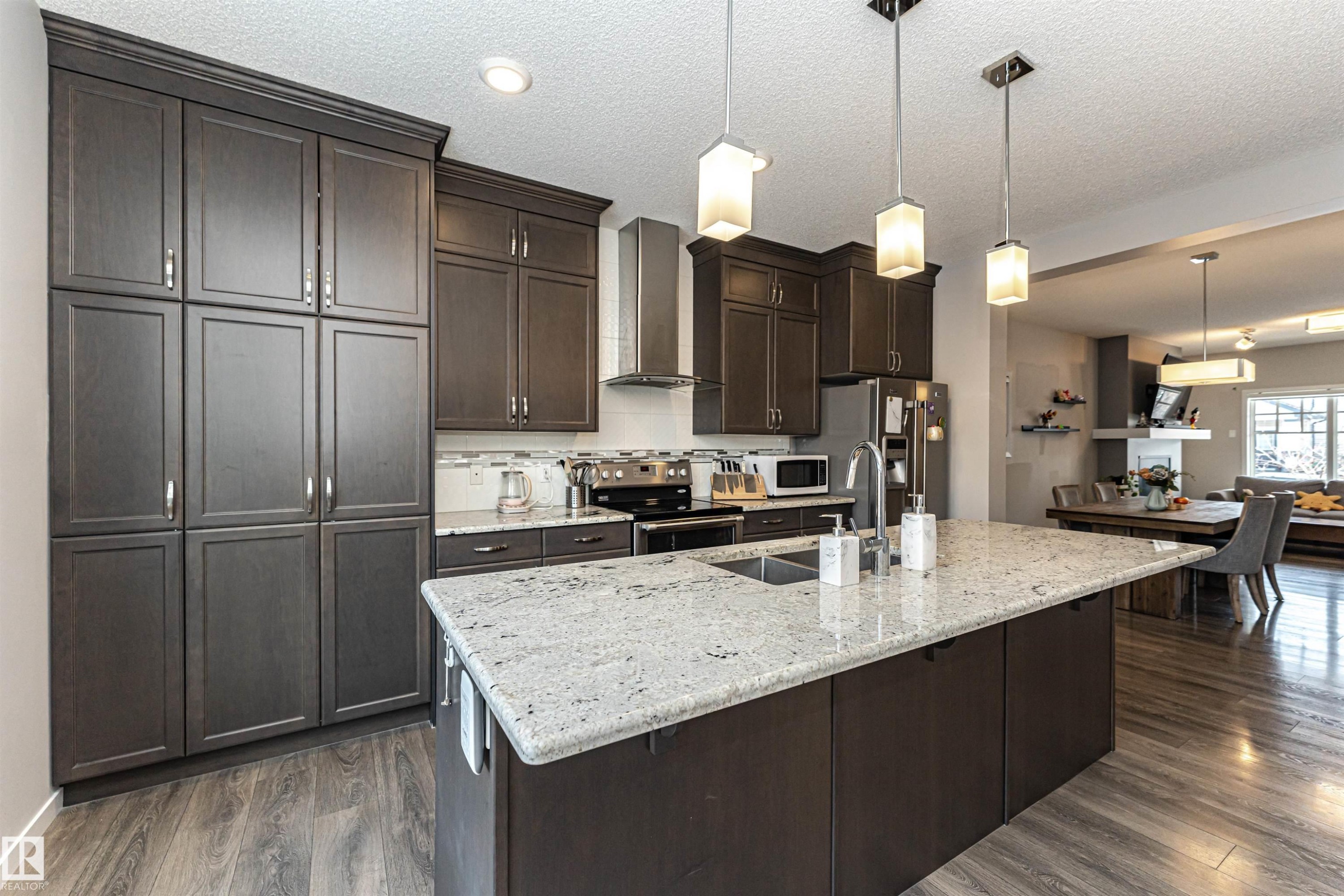 Kitchen with dark wood finish cabinetry, light stone counters, hanging light fixtures, an island with sink, and a textured ceiling - 4008 Allan Crescent, Edmonton, AB - Indoor Photo Showing Kitchen With Double Sink With Upgraded Kitchen