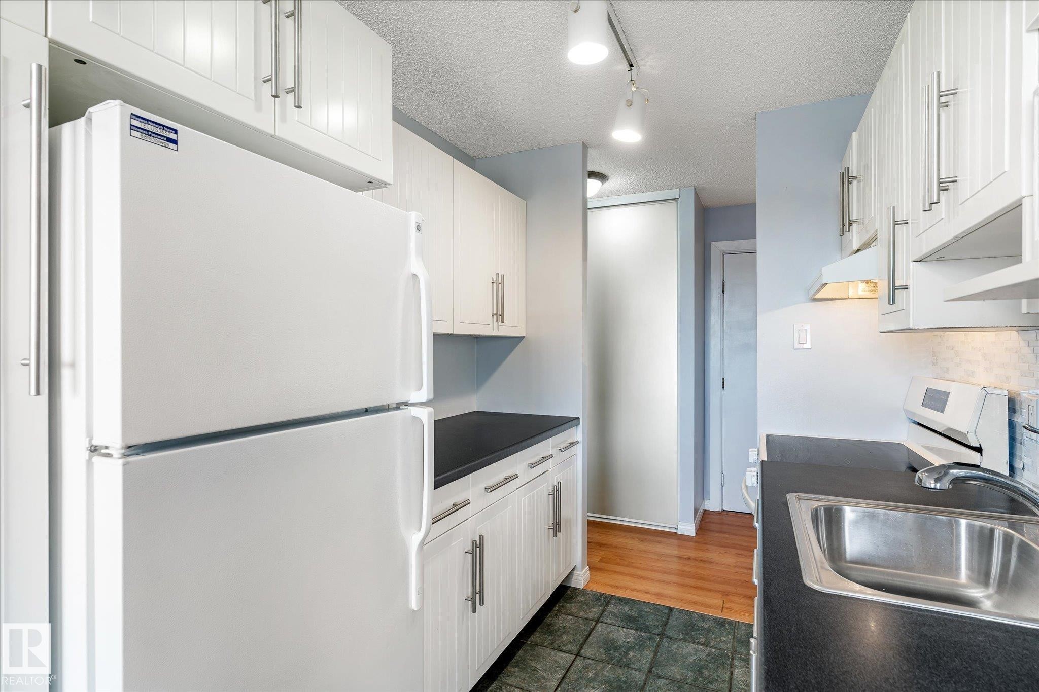 Kitchen featuring freestanding refrigerator, dark countertops, a textured ceiling, white cabinetry, and track lighting - 304 9725 106 Street, Edmonton, AB - Indoor Photo Showing Kitchen
