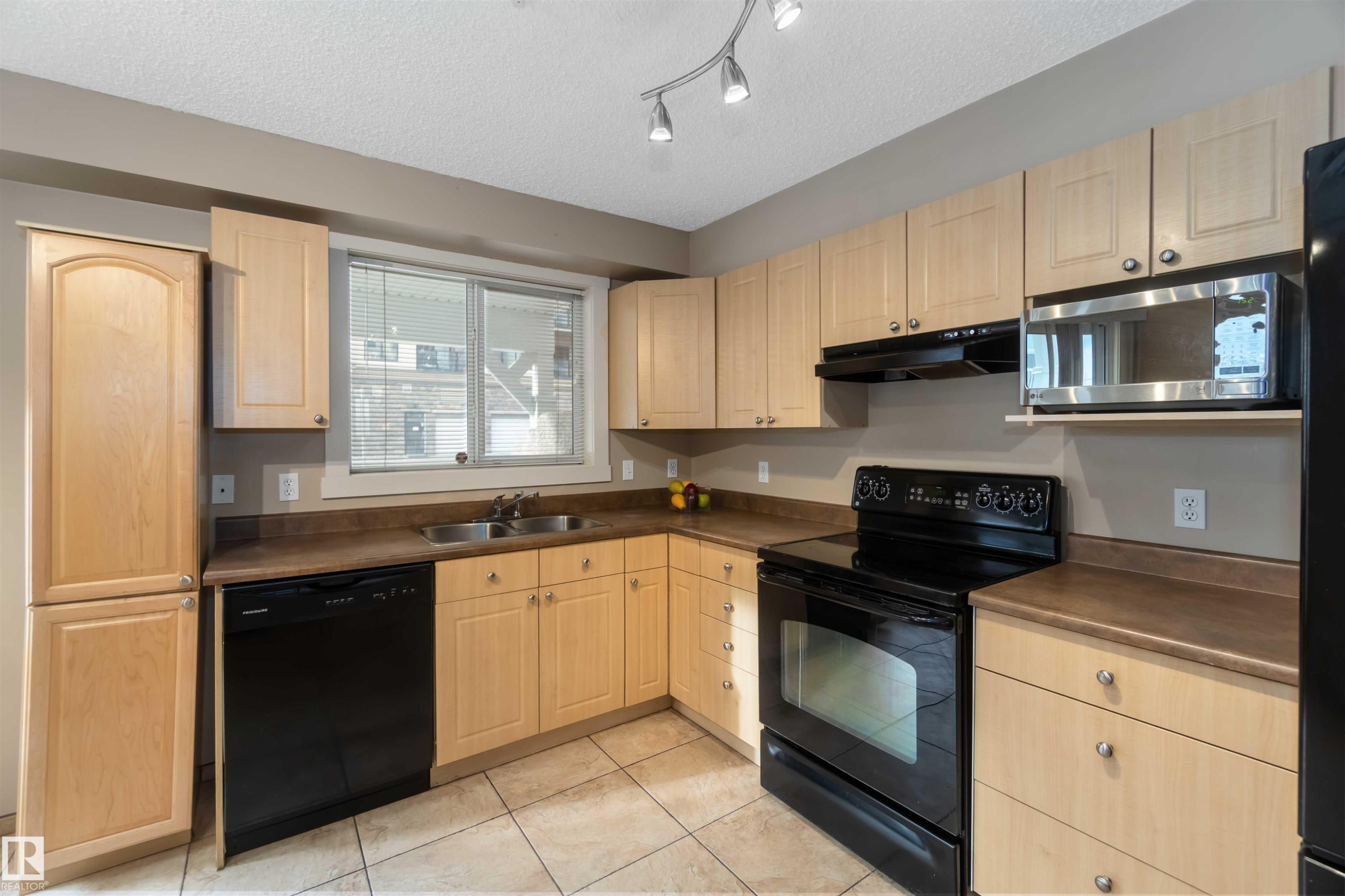 Kitchen with black appliances, light wood finish cabinetry, dark countertops, a textured ceiling, and light tile patterned floors - 113 10311 111 Street, Edmonton, AB - Indoor Photo Showing Kitchen With Double Sink