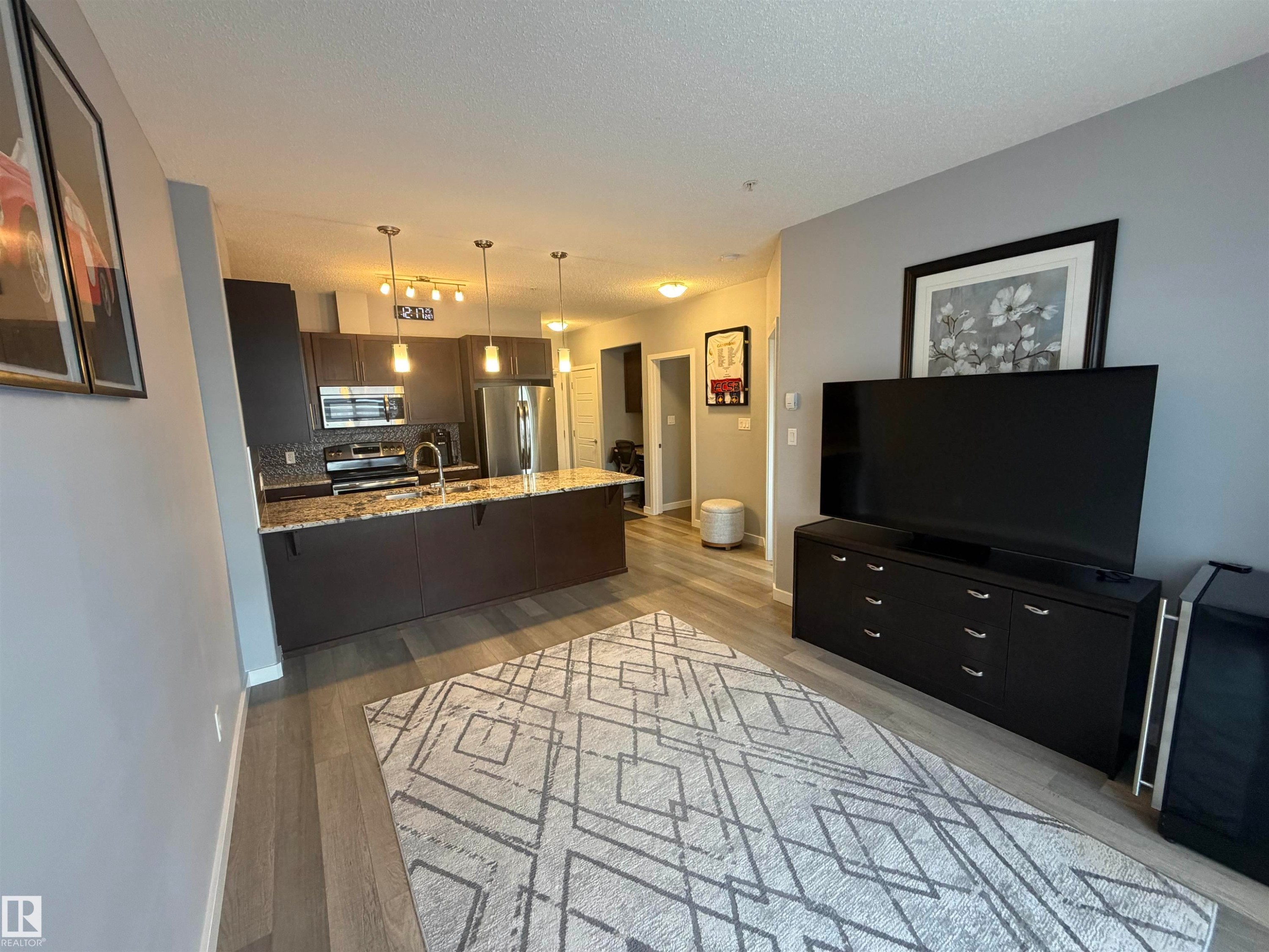 Kitchen with a peninsula, light stone counters, tasteful backsplash, stainless steel appliances, and hanging light fixtures - 417 1004 Rosenthal Boulevard, Edmonton, AB - Indoor Photo Showing Living Room