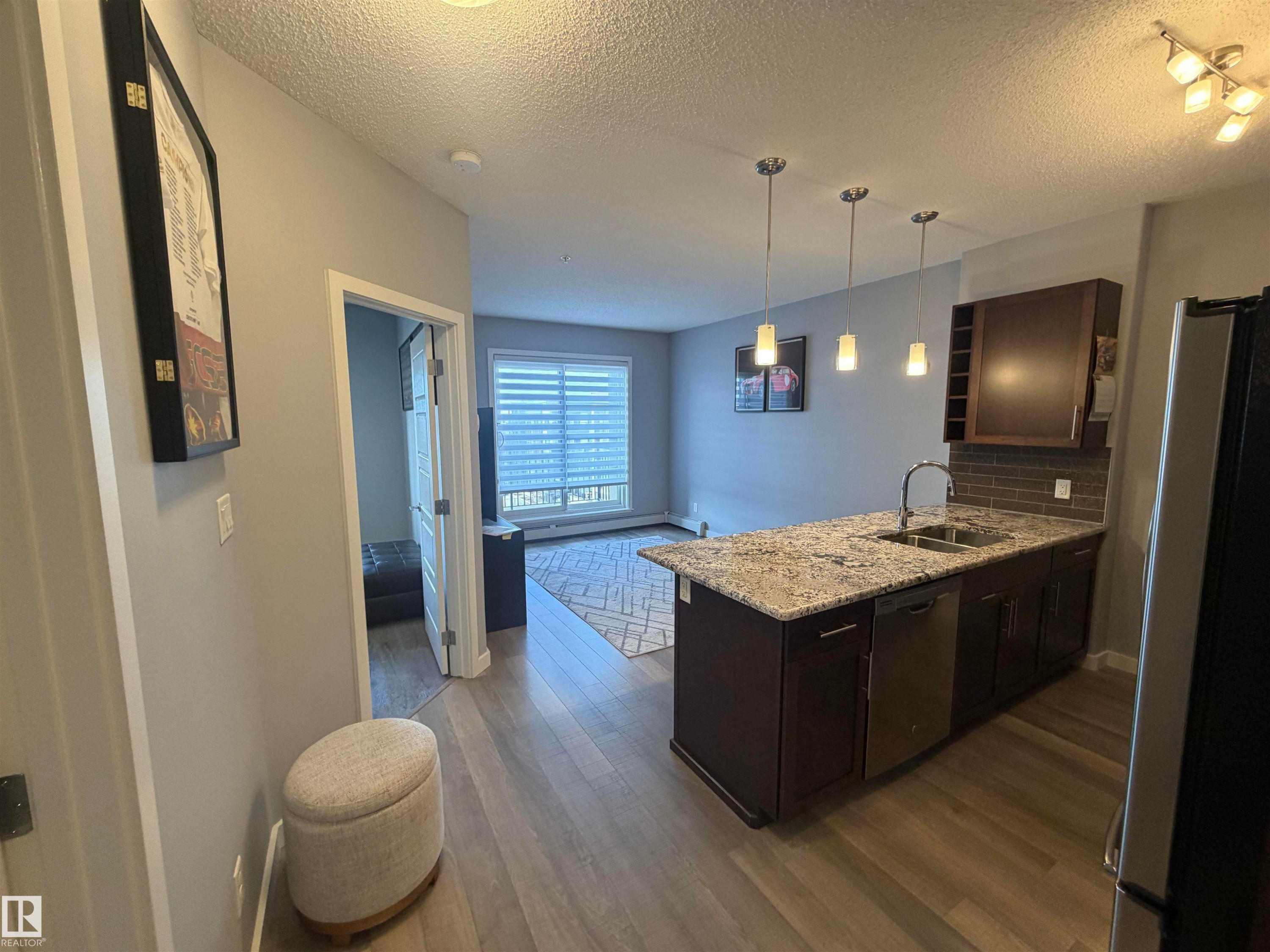 Kitchen with dark wood finish cabinetry, a peninsula, stainless steel appliances, light stone countertops, and dark wood-type flooring - 417 1004 Rosenthal Boulevard, Edmonton, AB - Indoor Photo Showing Kitchen With Double Sink