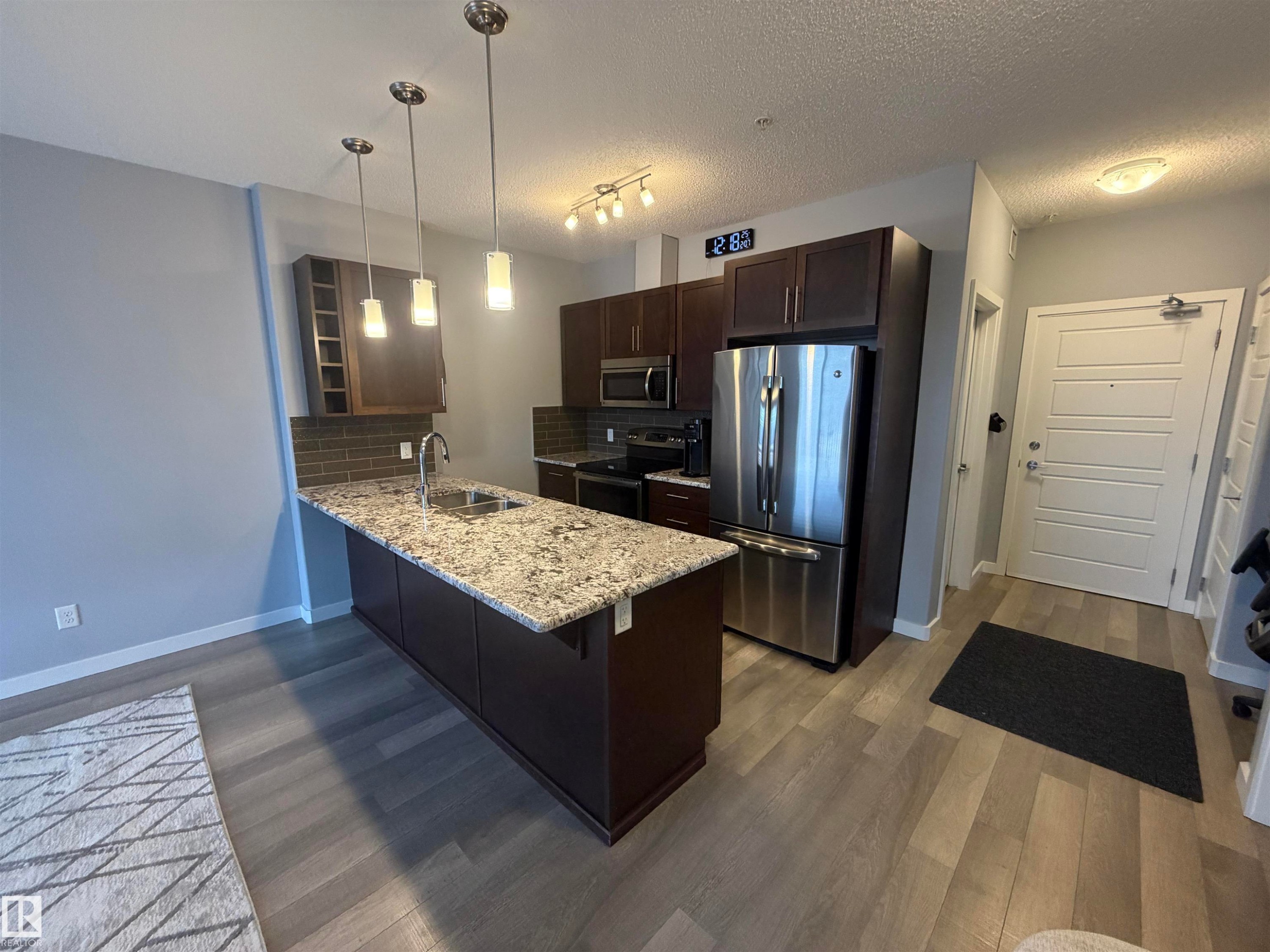 Kitchen with dark wood finish cabinets, stainless steel appliances, a peninsula, backsplash, and light stone countertops - 417 1004 Rosenthal Boulevard, Edmonton, AB - Indoor Photo Showing Kitchen With Stainless Steel Kitchen With Double Sink With Upgraded Kitchen