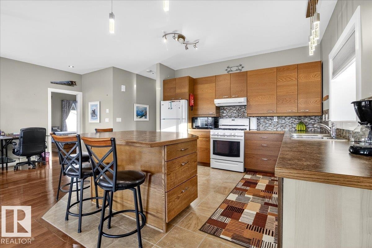 Kitchen with wood finish cabinets, white appliances, a kitchen breakfast bar, and hanging light fixtures - 5019 48 Avenue, Chipman, AB - Indoor Photo Showing Kitchen With Double Sink
