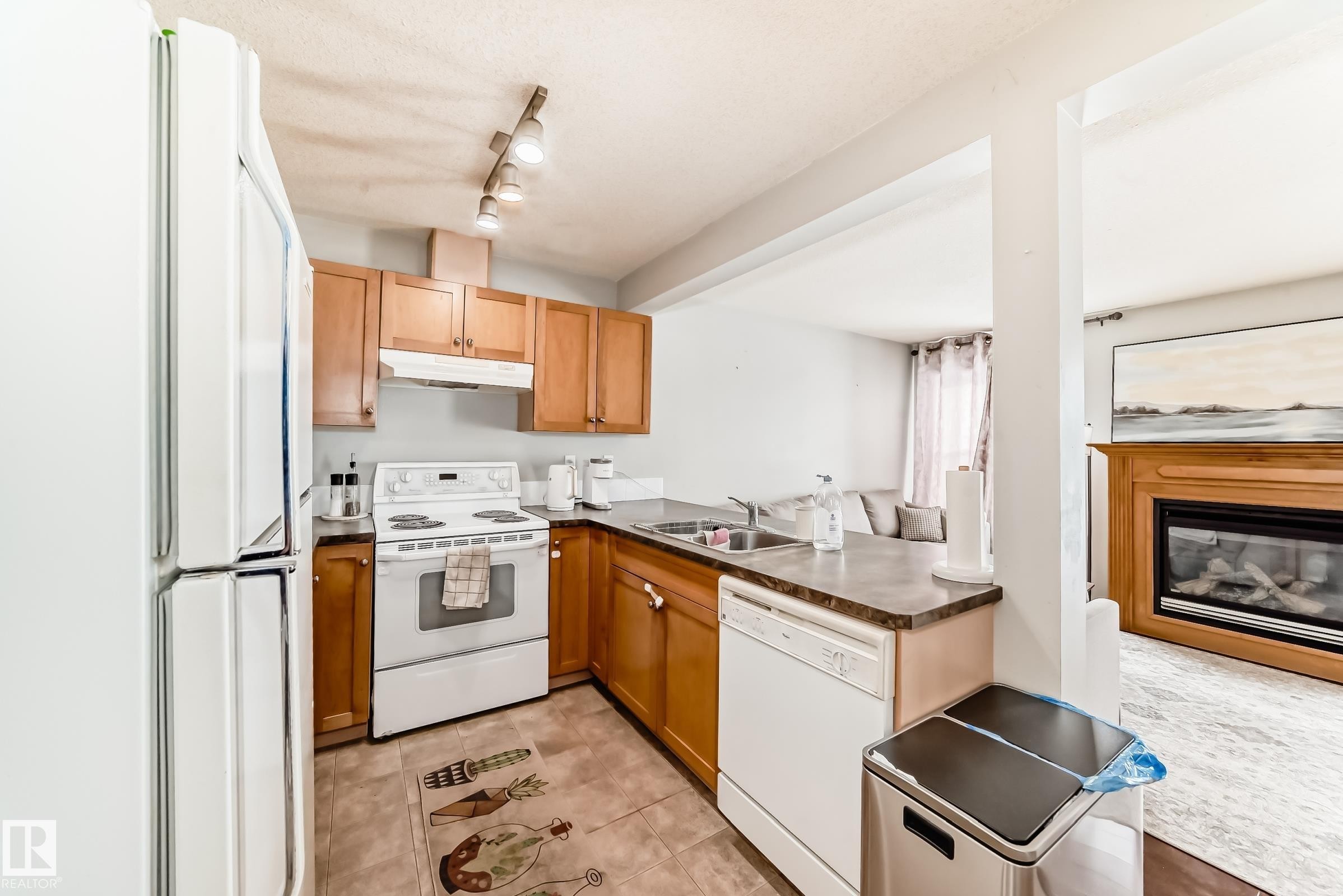 Kitchen featuring white appliances, a glass covered fireplace, a peninsula, rail lighting, and dark countertops - 185 230 Edwards Drive, Edmonton, AB