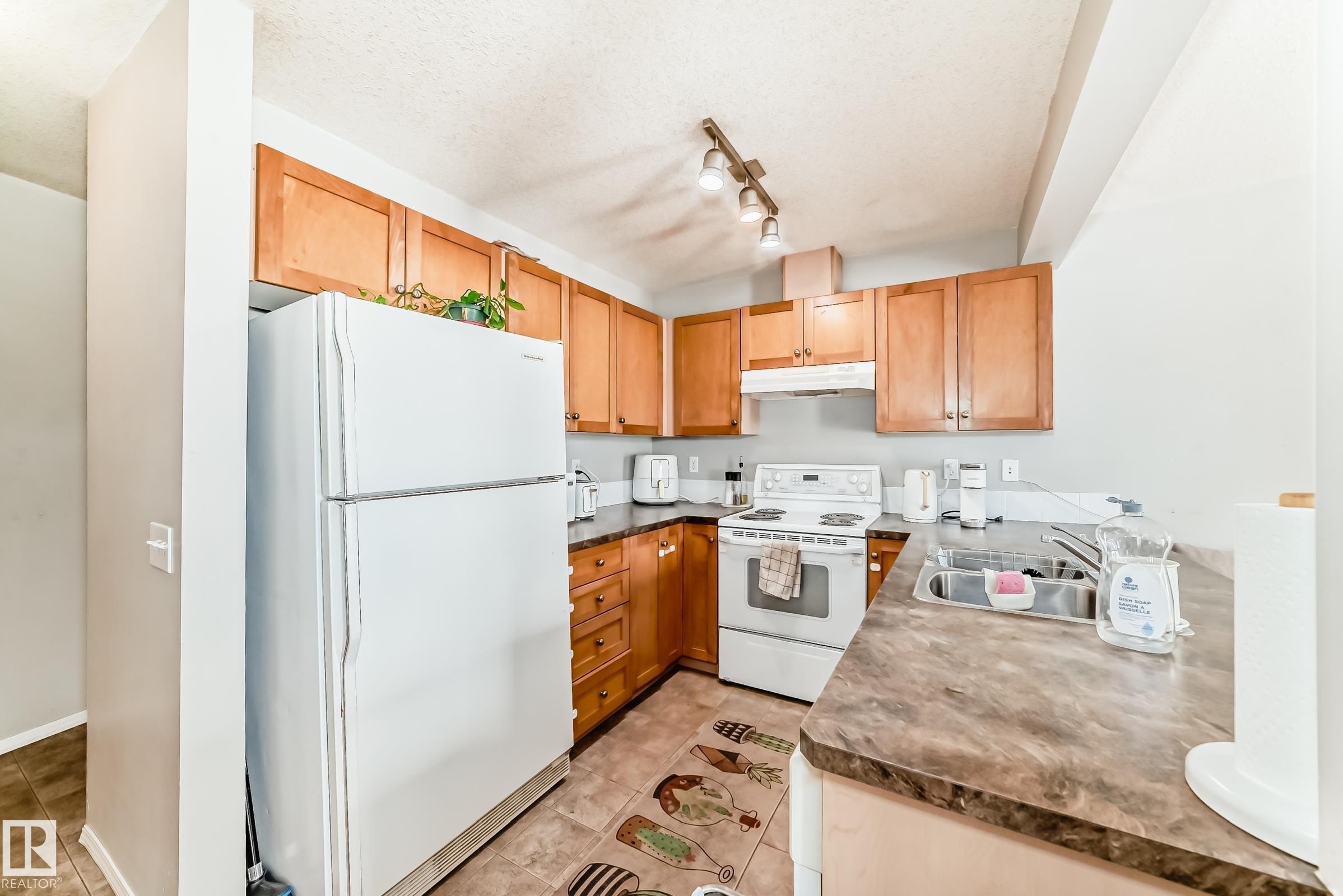 Kitchen with white appliances, rail lighting, dark countertops, light tile patterned floors, and wood finish cabinets - 185 230 Edwards Drive, Edmonton, AB