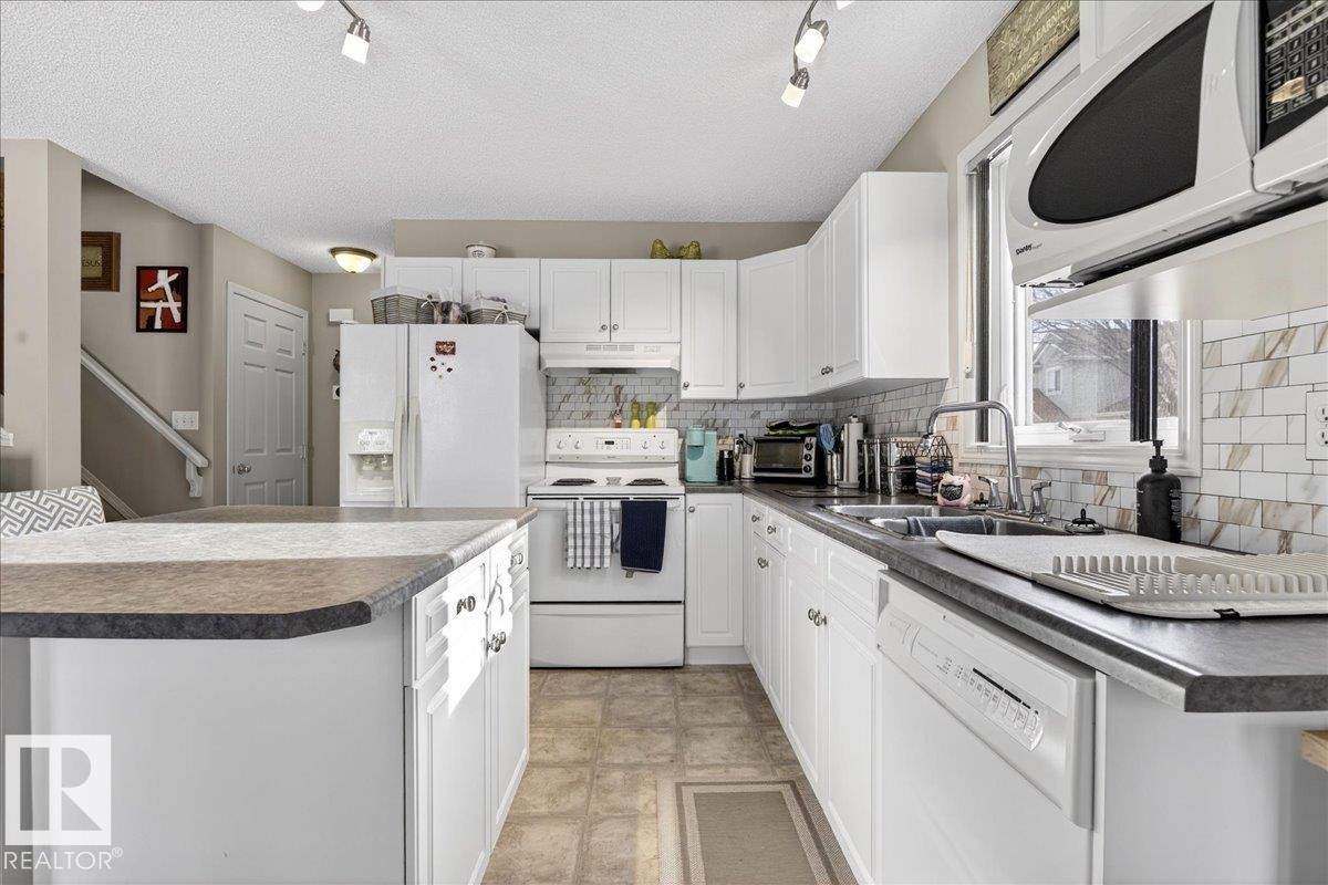 Kitchen featuring white appliances, dark countertops, white cabinetry, track lighting, and a kitchen island - 23 4020 21 Street, Edmonton, AB - Indoor Photo Showing Kitchen With Double Sink