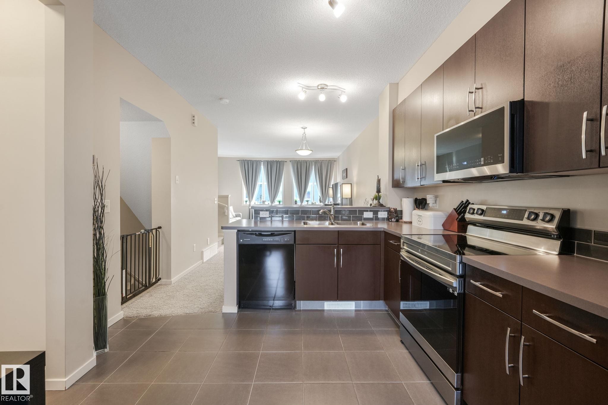Kitchen featuring stainless steel appliances, dark wood finish cabinetry, a peninsula, light tile patterned floors, and a textured ceiling - 91 603 Watt Boulevard, Edmonton, AB - Indoor Photo Showing Kitchen With Double Sink With Upgraded Kitchen
