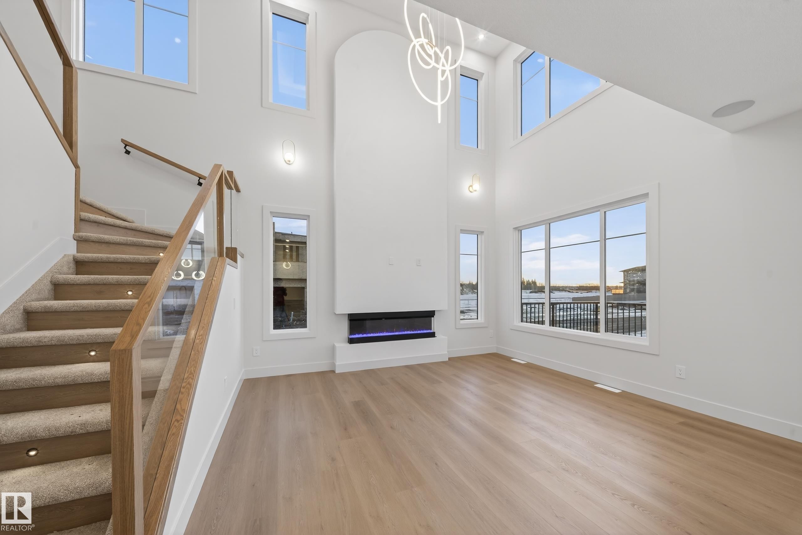 Unfurnished living room featuring light wood-style floors, a glass covered fireplace, hanging lights, and a high ceiling - 8924 Mayday Way, Edmonton, AB