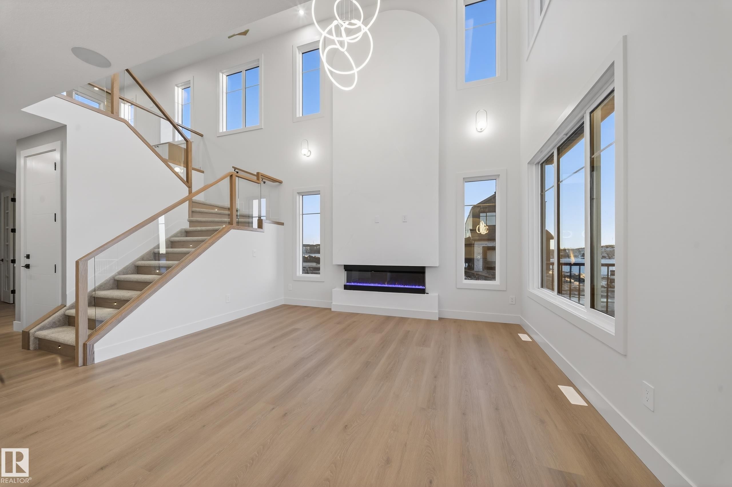 Unfurnished living room featuring light wood-type flooring, hanging lights, a glass covered fireplace, healthy amount of natural light, and a high ceiling - 8924 Mayday Way, Edmonton, AB