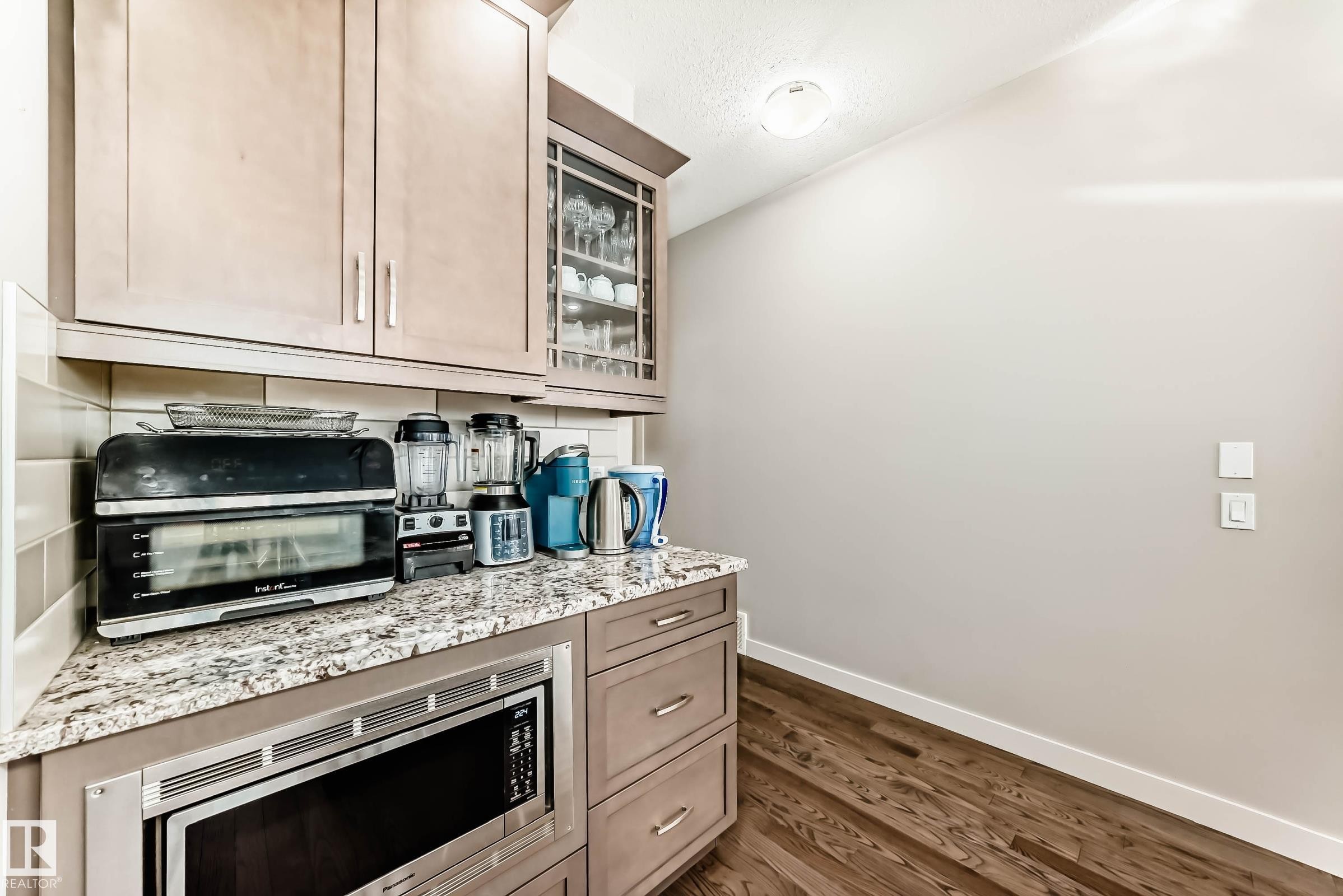 Kitchen featuring glass insert cabinets, stainless steel microwave, dark wood finished floors, and light stone counters - 7719 Getty Wynd, Edmonton, AB - Indoor Photo Showing Kitchen