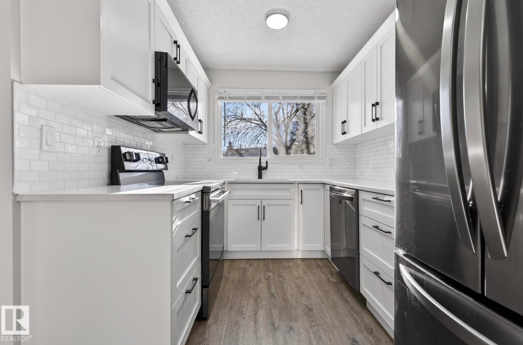 Kitchen featuring stainless steel appliances, dark wood-type flooring, white cabinets, a textured ceiling, and light stone counters - 9969 171 Ave, Edmonton, AB - Indoor Photo Showing Kitchen With Upgraded Kitchen