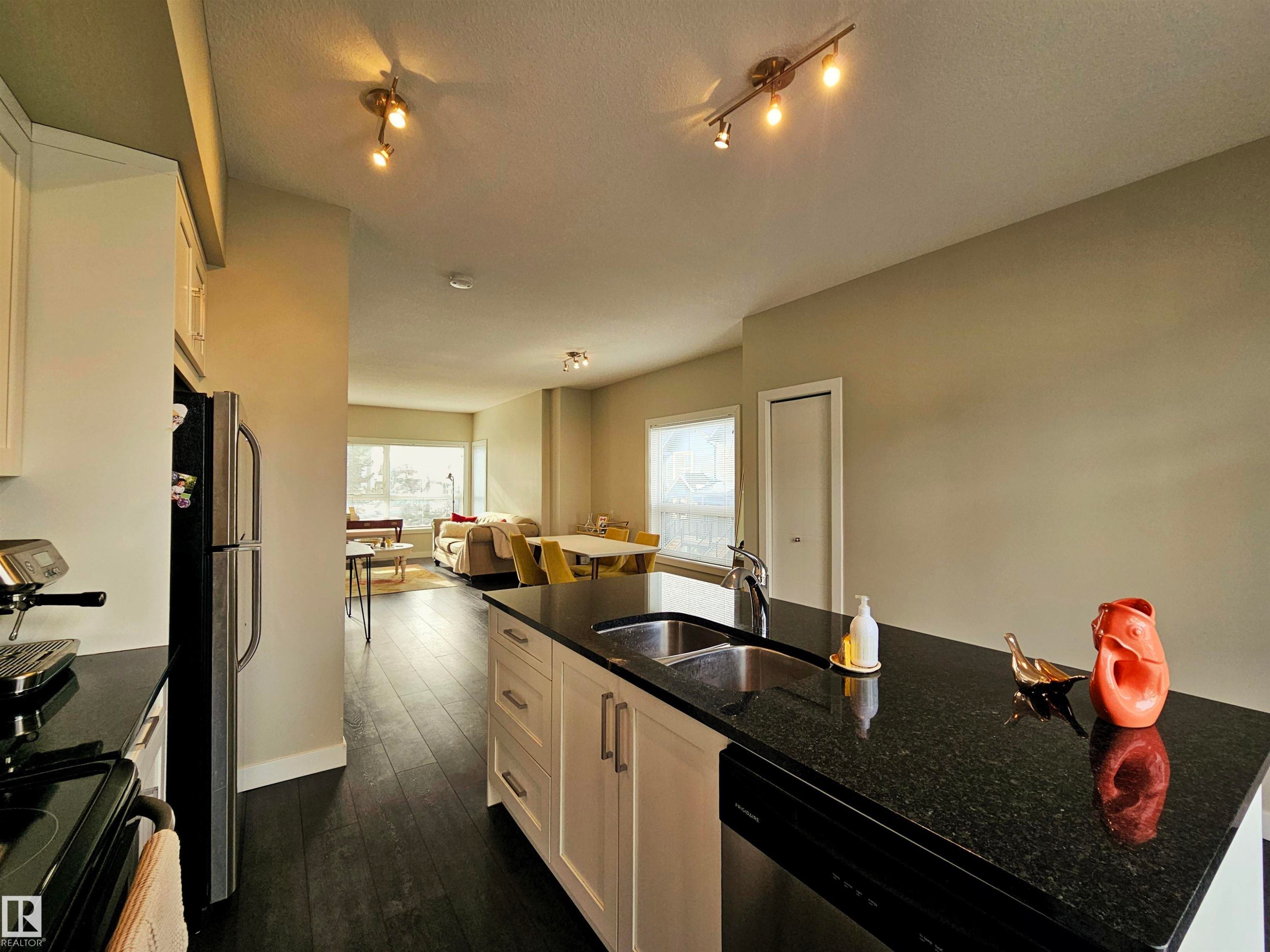 Kitchen featuring dark stone countertops, a center island with sink, dark wood-style flooring, white cabinetry, and stainless steel appliances - 129 2560 Pegasus Boulevard, Edmonton, AB - Indoor Photo Showing Kitchen With Double Sink