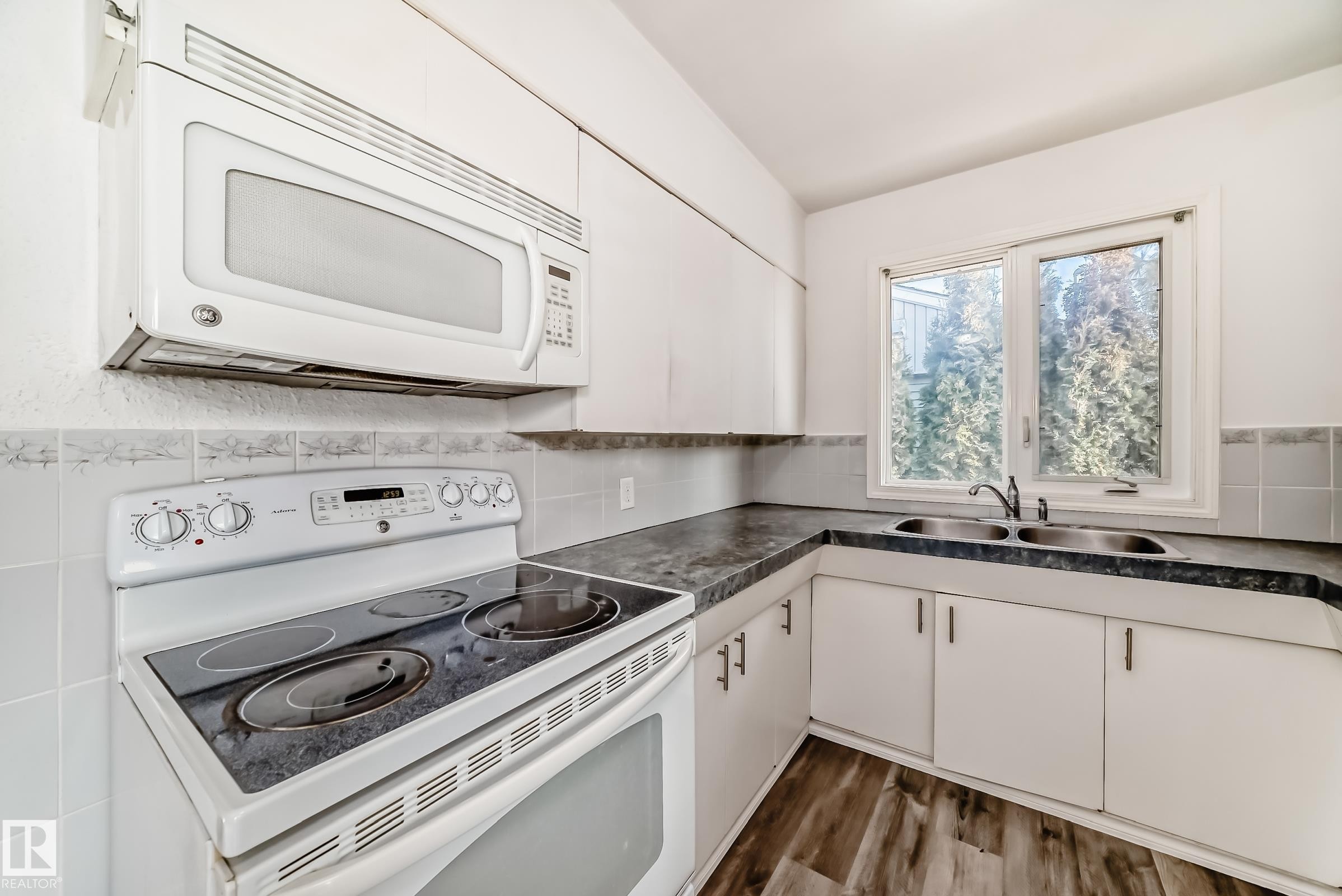 Kitchen featuring white appliances, white cabinets, dark countertops, and wood finished floors - 8639 64 Avenue Nw, Edmonton, AB - Indoor Photo Showing Kitchen With Double Sink