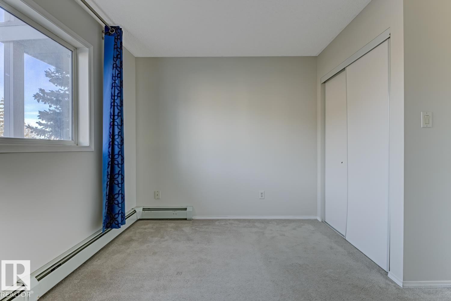This room features carpeted flooring, a window with a blue curtain, baseboard heating, and a white closet with sliding doors - 237 17447 98A Avenue, Edmonton, AB - Indoor Photo Showing Other Room