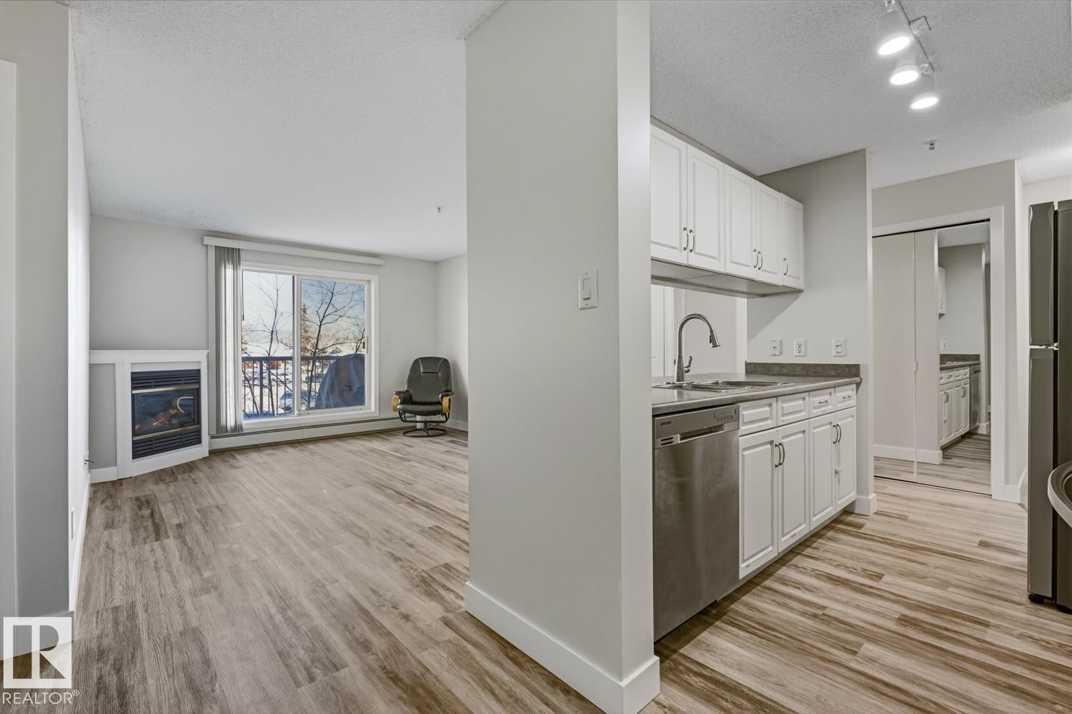 Kitchen with white cabinets, light wood-style flooring, a textured ceiling, stainless steel dishwasher, and a glass covered fireplace - 237 17447 98A Avenue, Edmonton, AB - Indoor Photo Showing Kitchen With Fireplace