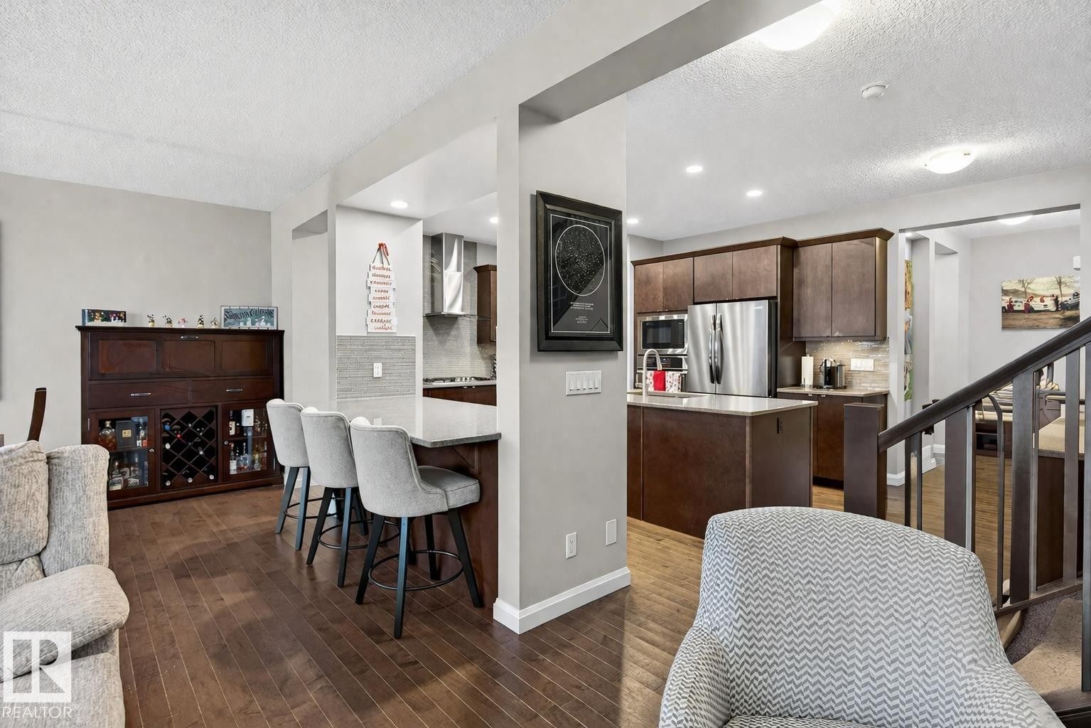 The open concept living area features hardwood floors, a kitchen with dark cabinetry and stainless steel appliances, and a breakfast bar with seating - 17838 60A Street, Edmonton, AB - Indoor