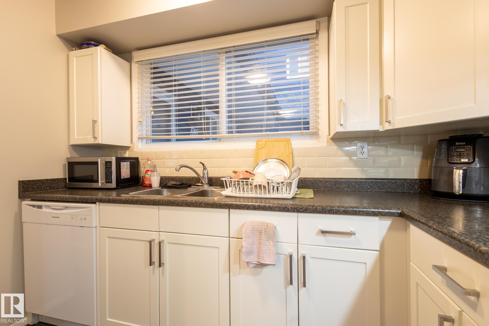 Kitchen with dark countertops, white dishwasher, stainless steel microwave, white cabinets, and tasteful backsplash - 3530 42 Street, Edmonton, AB - Indoor Photo Showing Kitchen With Double Sink