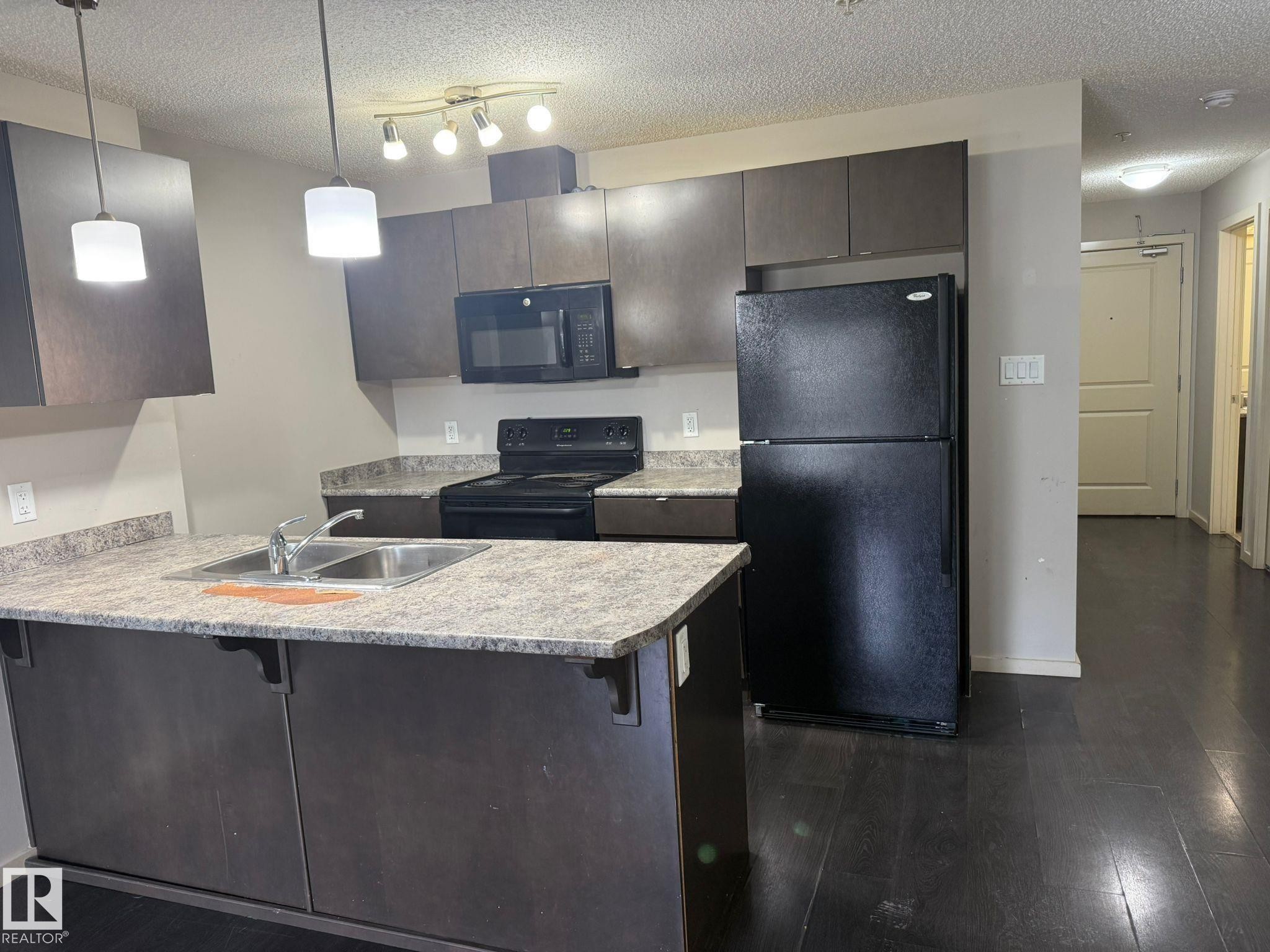 Kitchen with a breakfast bar area, black appliances, a textured ceiling, light countertops, and dark wood finished floors - 211 340 Windermere Road, Edmonton, AB - Indoor Photo Showing Kitchen With Double Sink