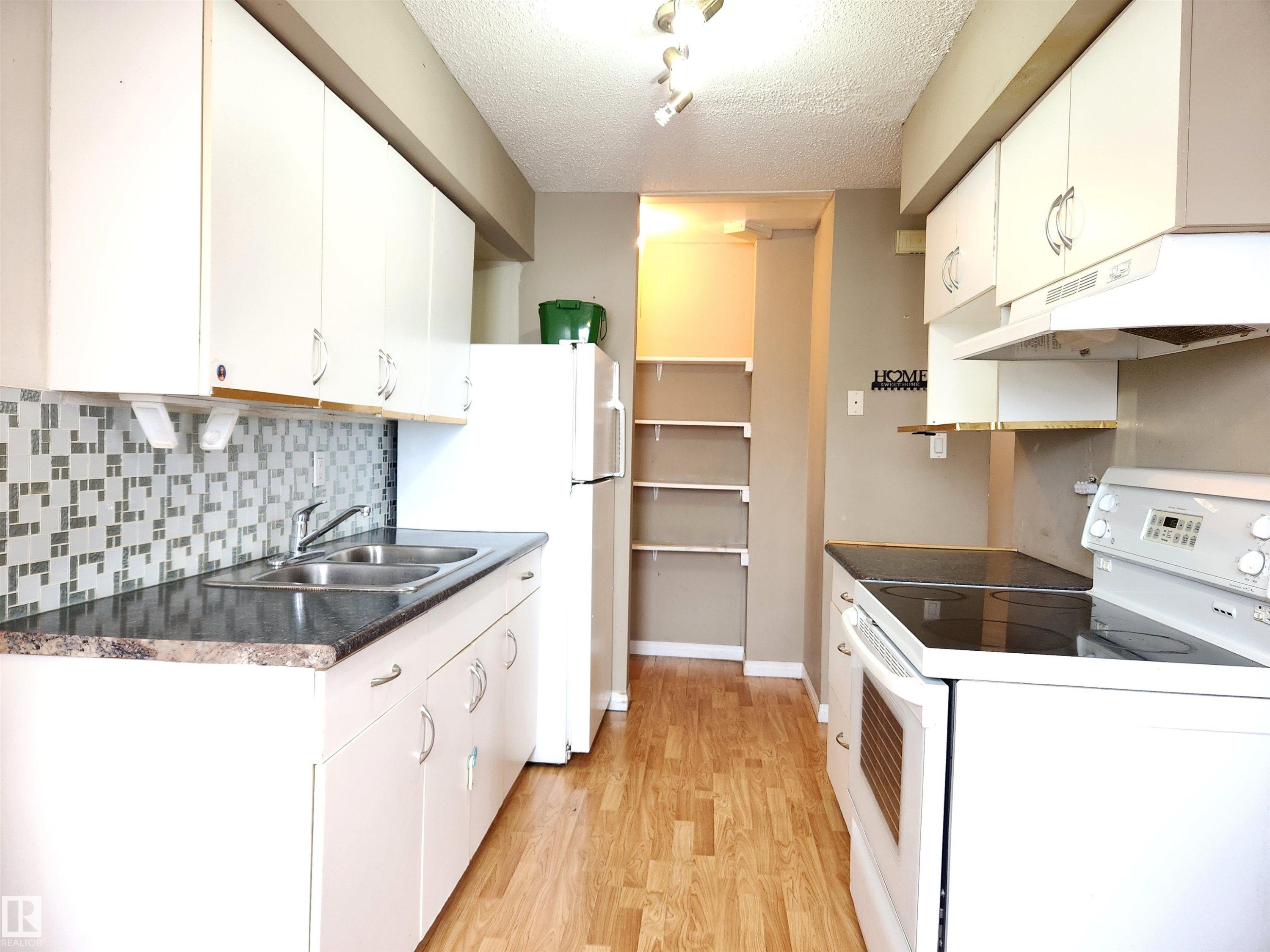 Kitchen with white appliances, a textured ceiling, white cabinetry, light wood finished floors, and dark countertops - 6 14320 80 Street, Edmonton, AB - Indoor Photo Showing Kitchen With Double Sink
