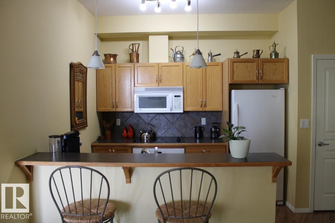 Kitchen with a breakfast bar, white appliances, pendant lighting, dark countertops, and decorative backsplash - #330 4831 104A St Nw, Edmonton, AB - Indoor Photo Showing Kitchen