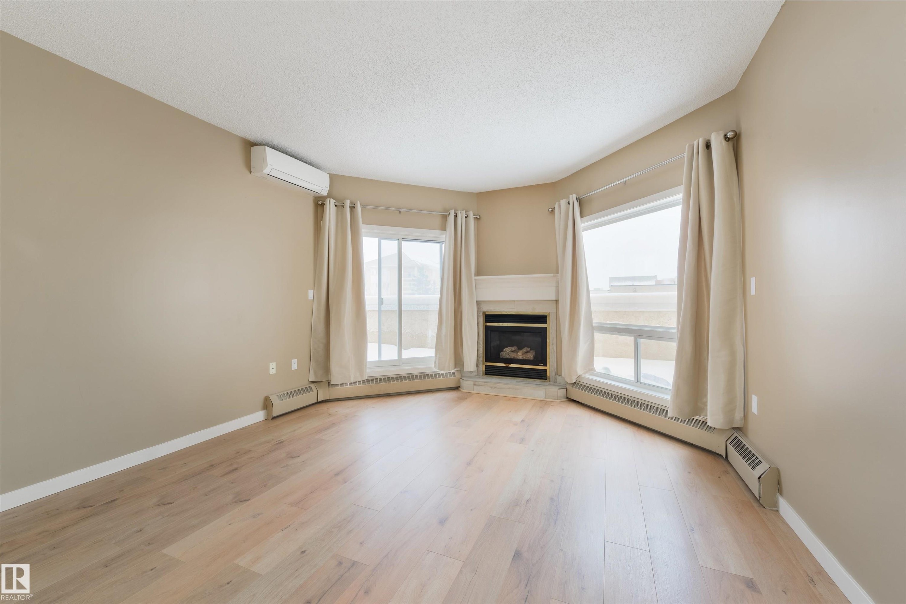 Unfurnished living room featuring a glass covered fireplace, light wood-style flooring, a baseboard heating unit, a baseboard radiator, and healthy amount of natural light - 232 15499 Castle Downs Road, Edmonton, AB - Indoor Photo Showing Living Room With Fireplace