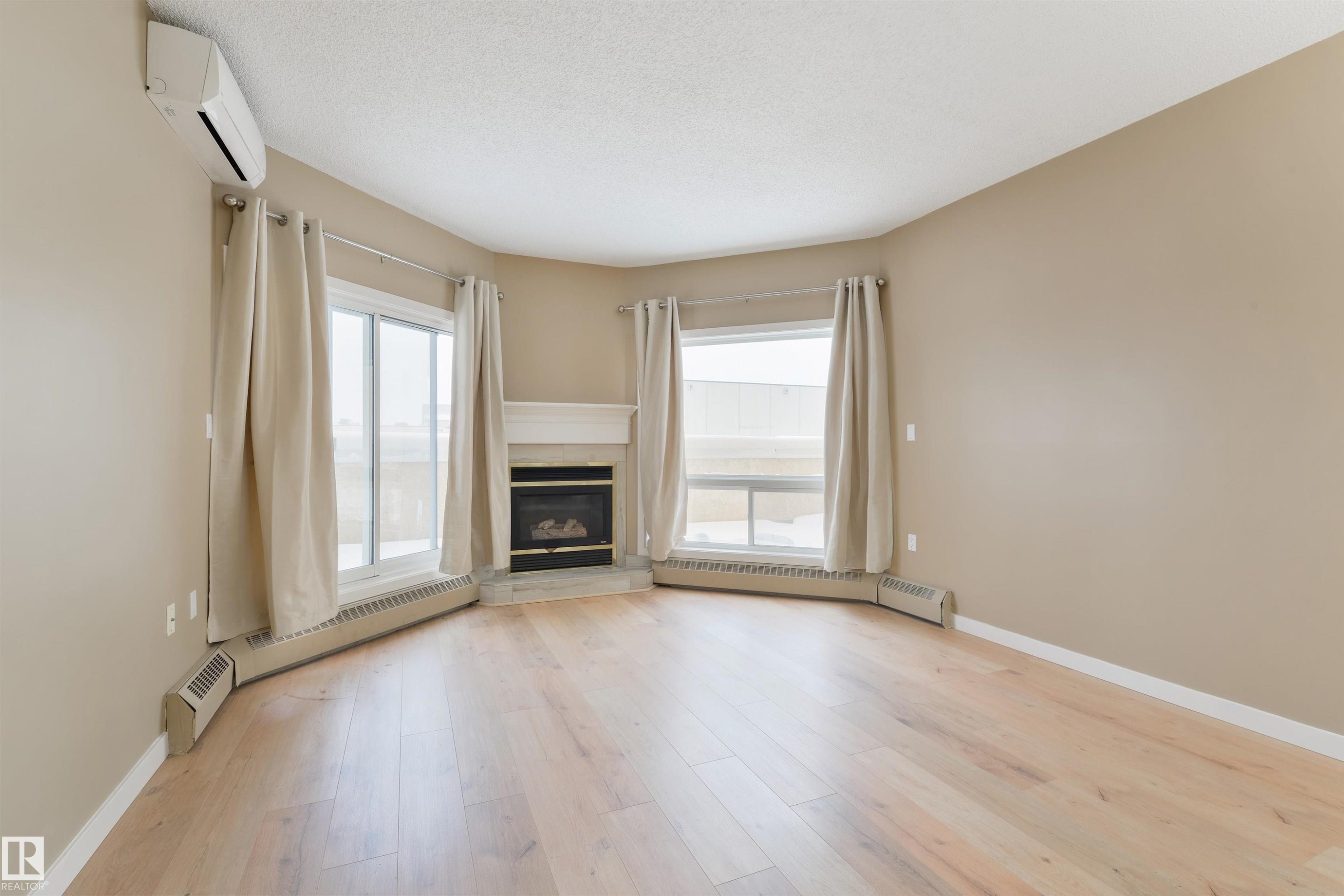 Unfurnished living room featuring a glass covered fireplace, a baseboard heating unit, a baseboard radiator, light wood finished floors, and healthy amount of natural light - 232 15499 Castle Downs Road, Edmonton, AB - Indoor Photo Showing Other Room With Fireplace
