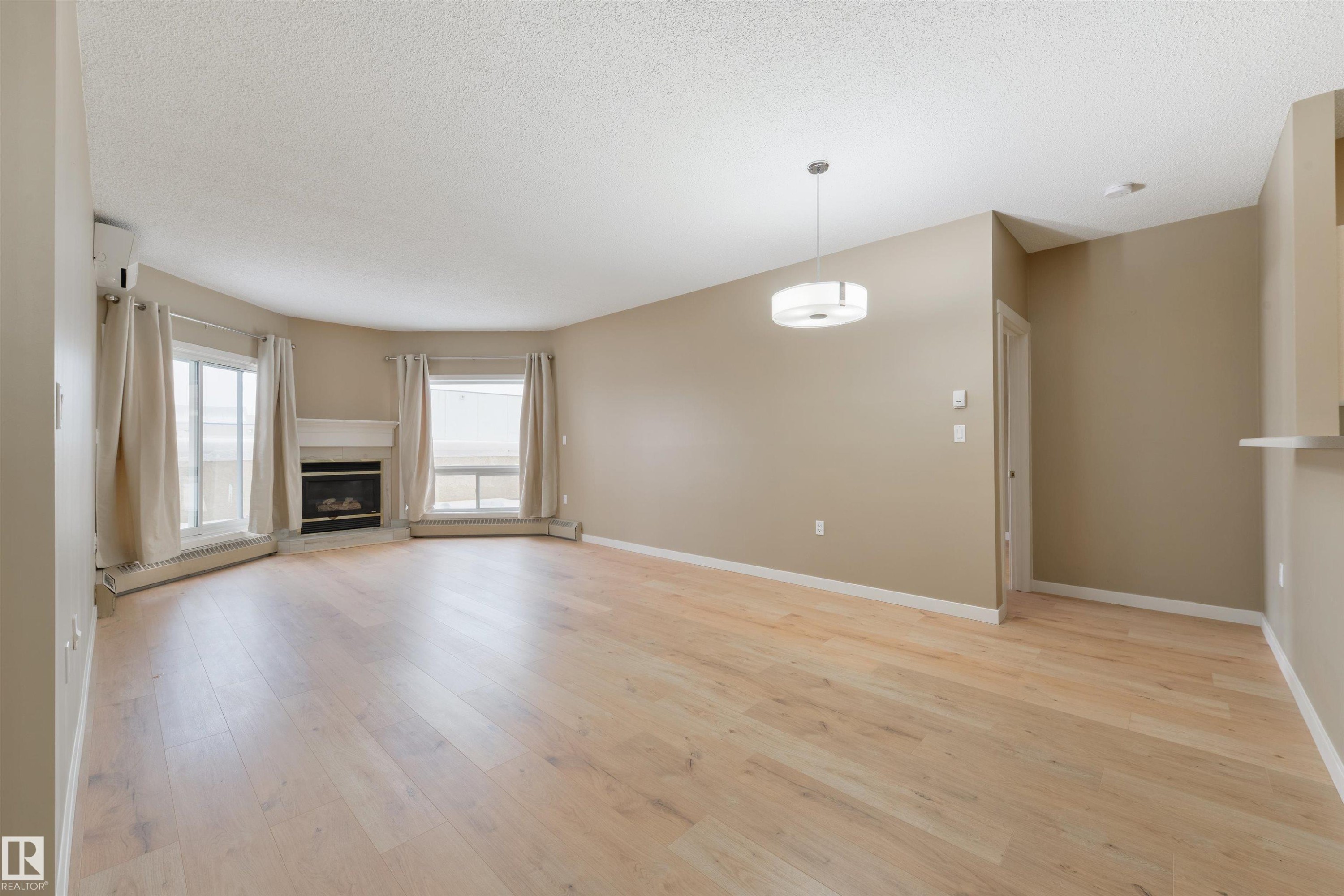 Unfurnished living room with light wood finished floors, a textured ceiling, a fireplace, and a baseboard heating unit - 232 15499 Castle Downs Road, Edmonton, AB - Indoor Photo Showing Living Room With Fireplace